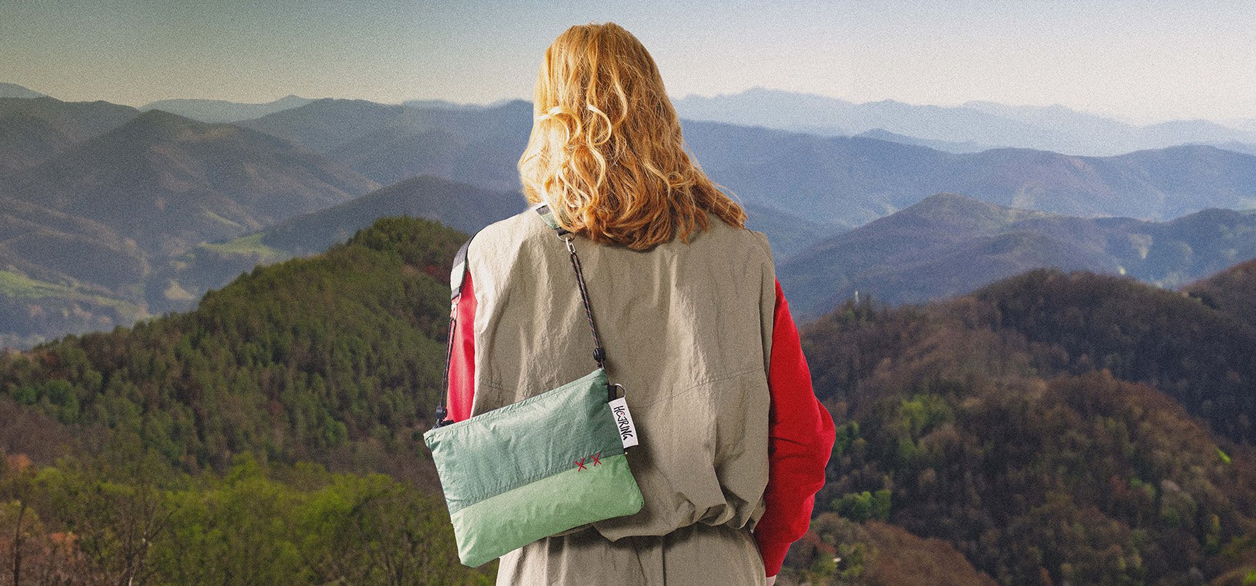 Person with curly blonde hair standing on a hilltop overlooking mountains during daytime, wearing a beige jacket with red sleeves and carrying a green crossbody bag.