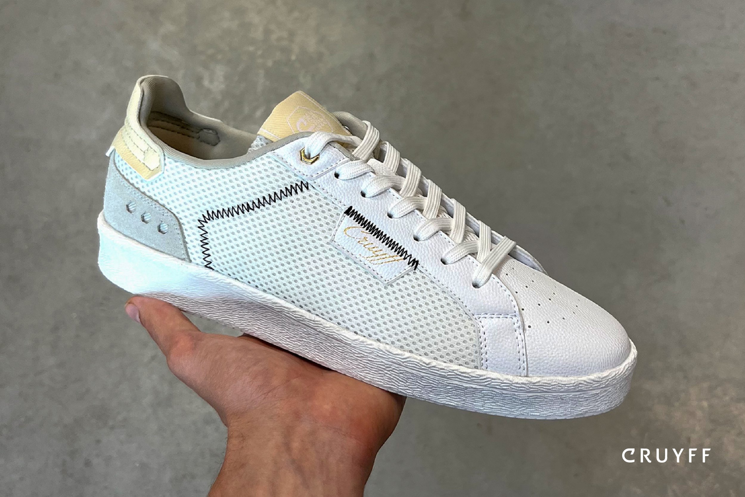 A white athletic sneaker with mesh and leather details, held in a person's hand against a plain background. The shoe has the brand name 'Cruyff' on a fabric tag on its side.