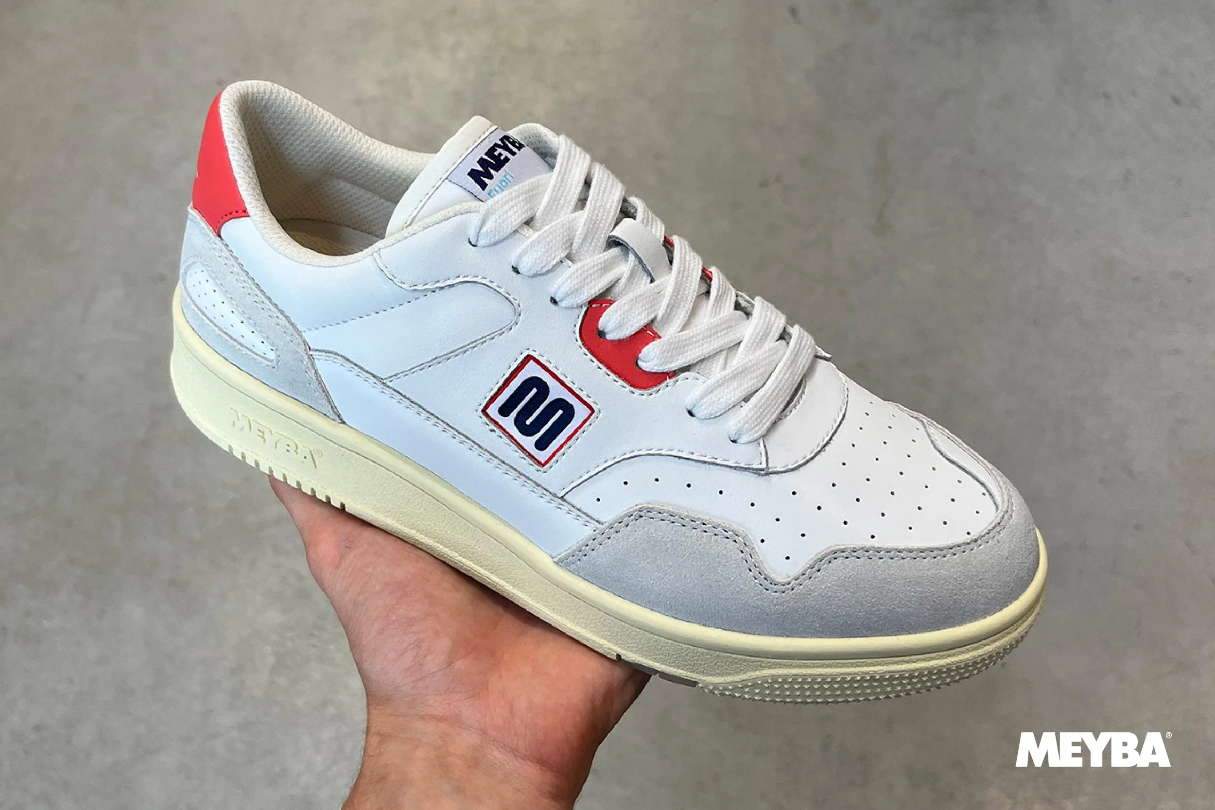White and grey sneaker with red and navy logo details, being held in a hand against a grey background, with the 'MEYBA' brand visible on the sole and tongue.