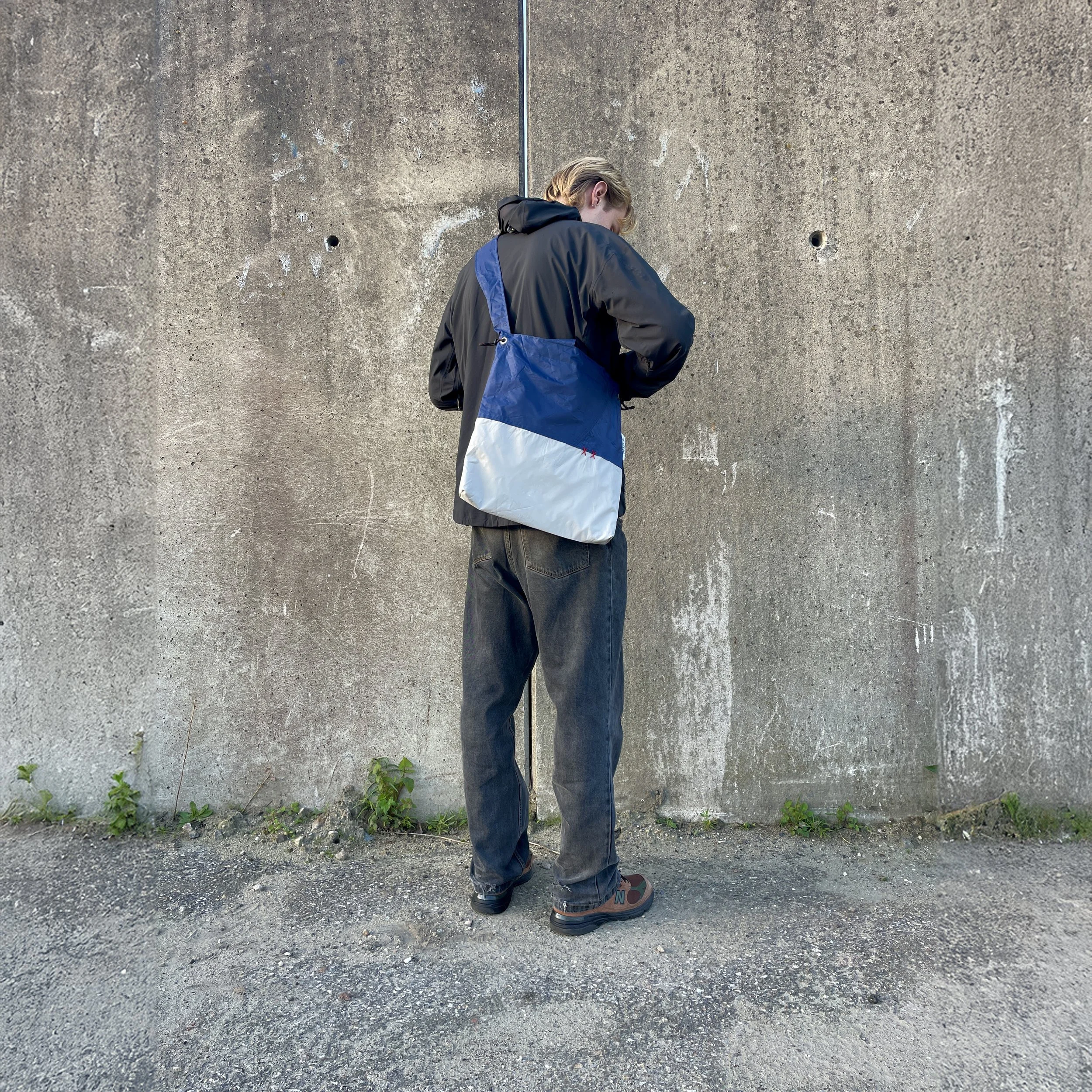A person standing outdoors against a concrete wall, wearing a black jacket, dark jeans, hiking shoes, and carrying a blue and white backpack.