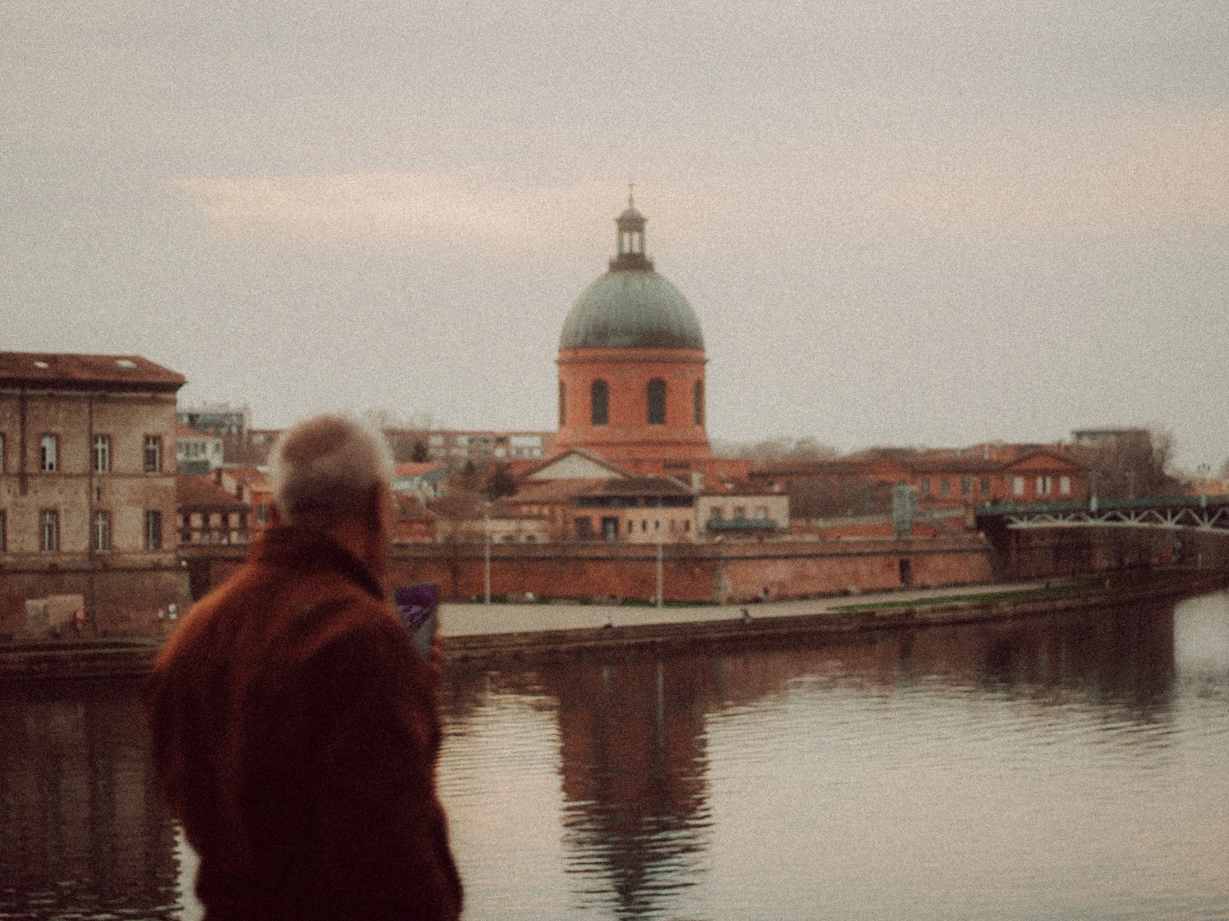Un homme regarde une rivière avec un bâtiment à dôme en arrière-plan, probablement à Toulouse, France.