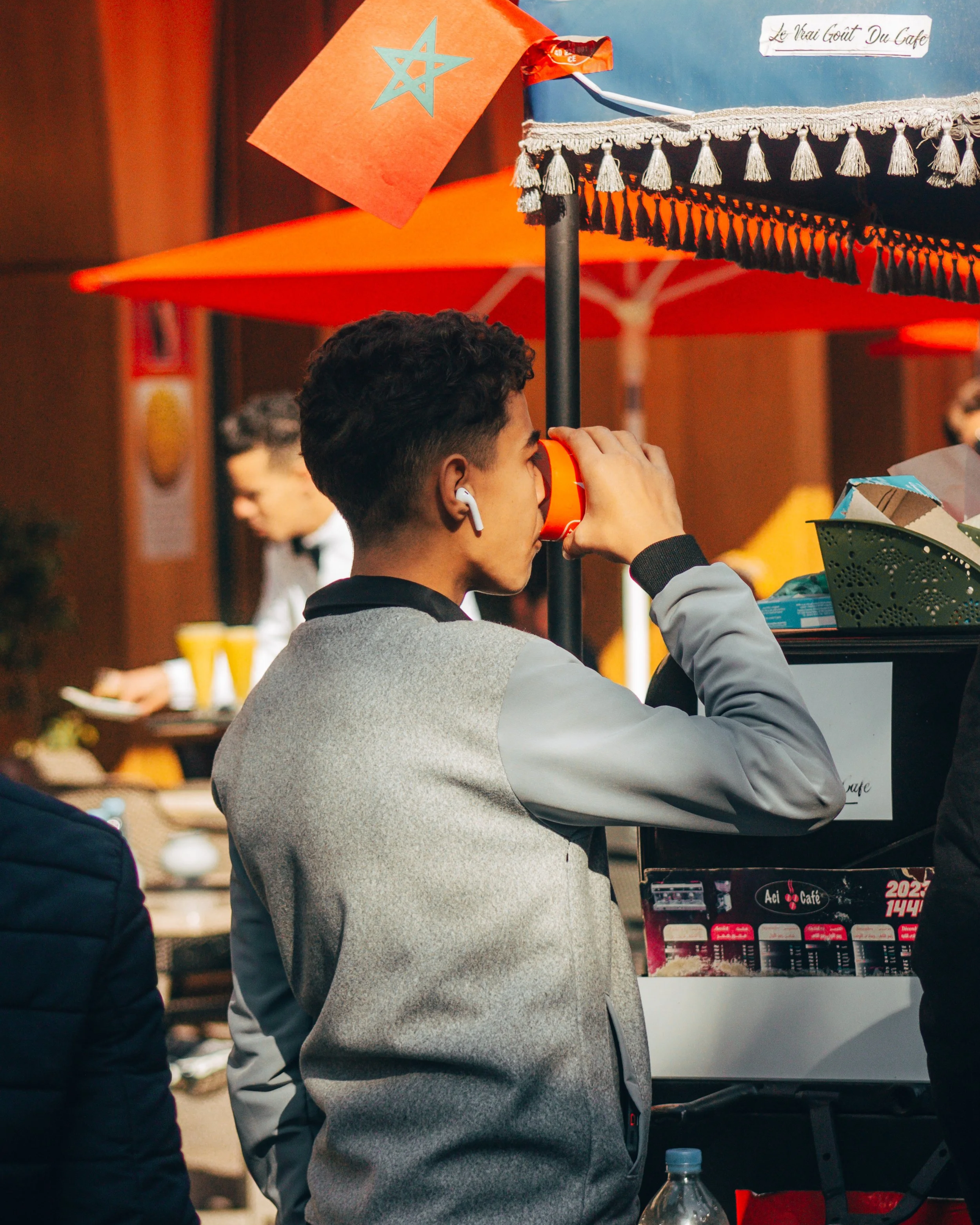 Jeune homme en train de boire dans une tasse en se tenant près d'un stand, avec des parasols orange en arrière-plan et une petite drapeau orange avec une étoile bleue