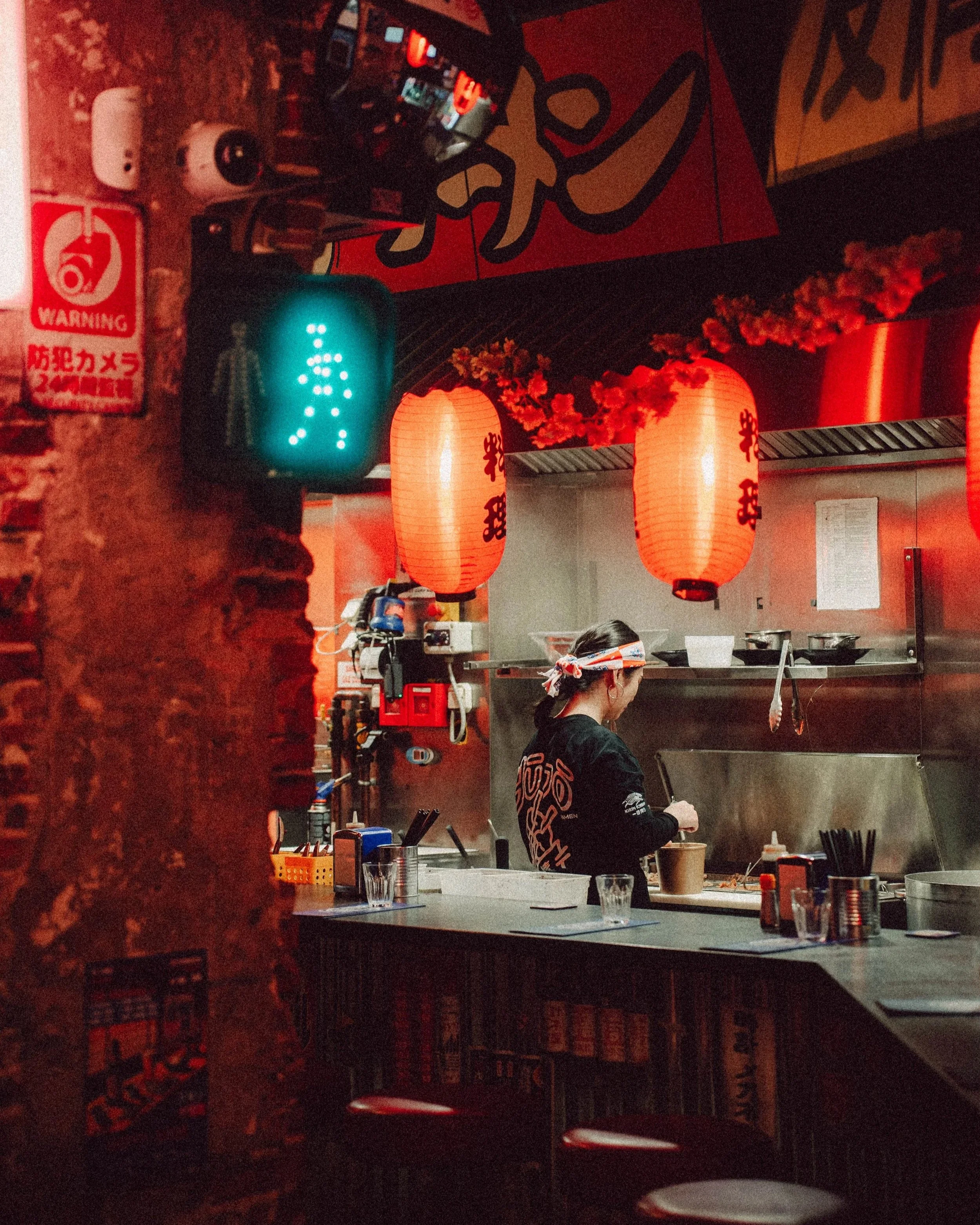 Intérieur d'un restaurant japonais avec décor en rouge, lanternes japonaises, une femme travaillant derrière le comptoir, ambiance chaleureuse et éclairage tamisé.