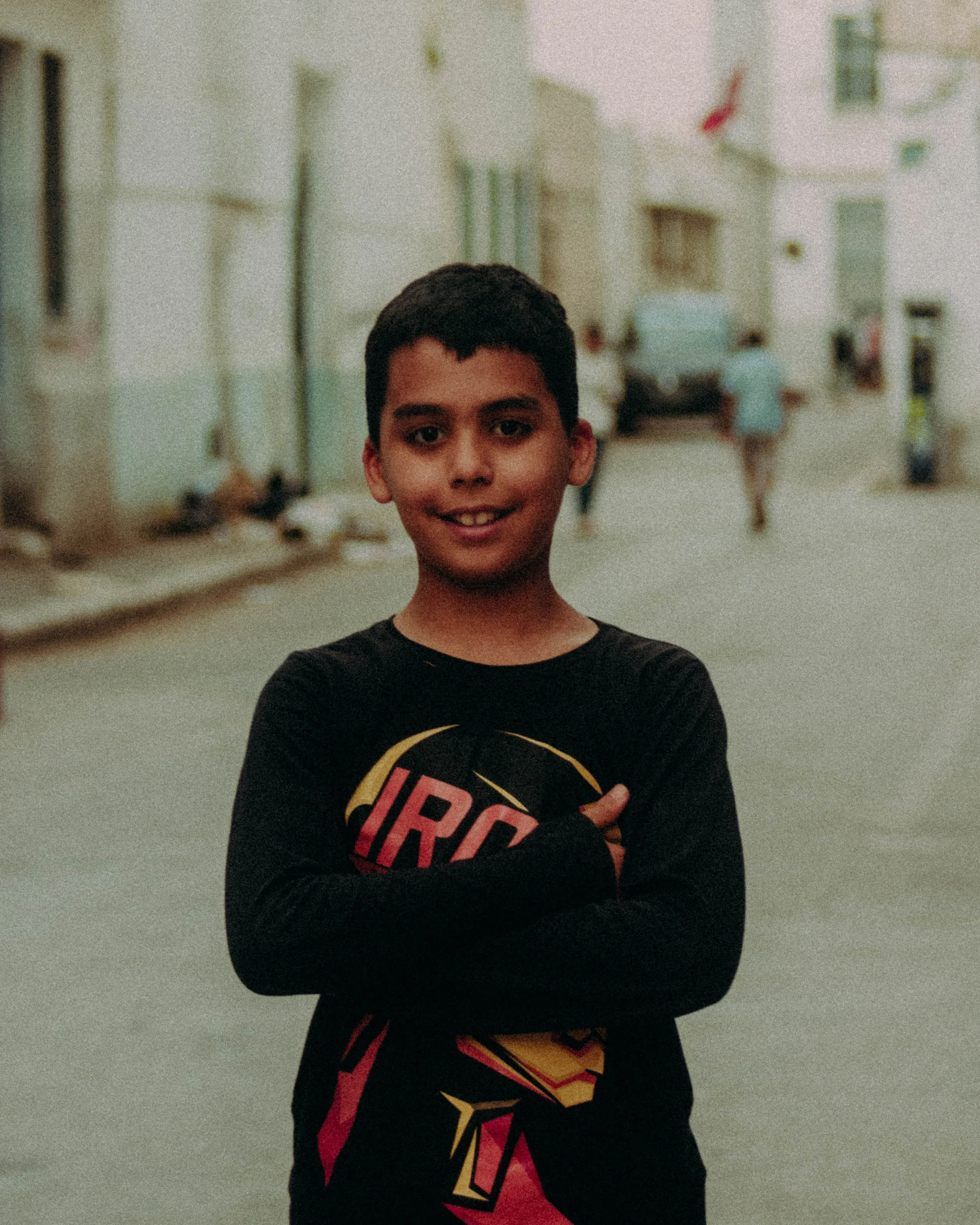 Un garçon souriant portant un t-shirt noir avec des bras croisés, debout dans une rue.
