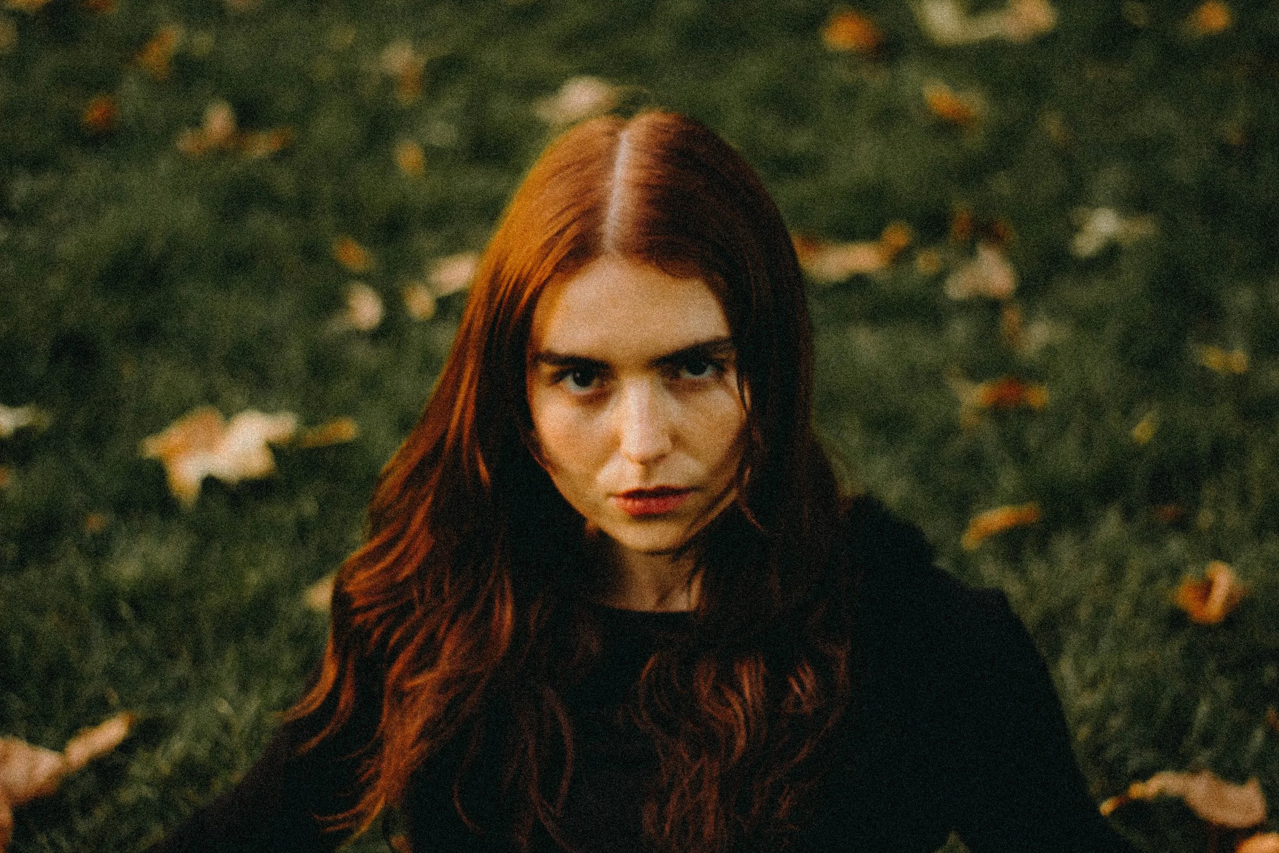 Une femme rousse avec un regard intense, assise sur de l'herbe avec des feuilles d'automne autour.