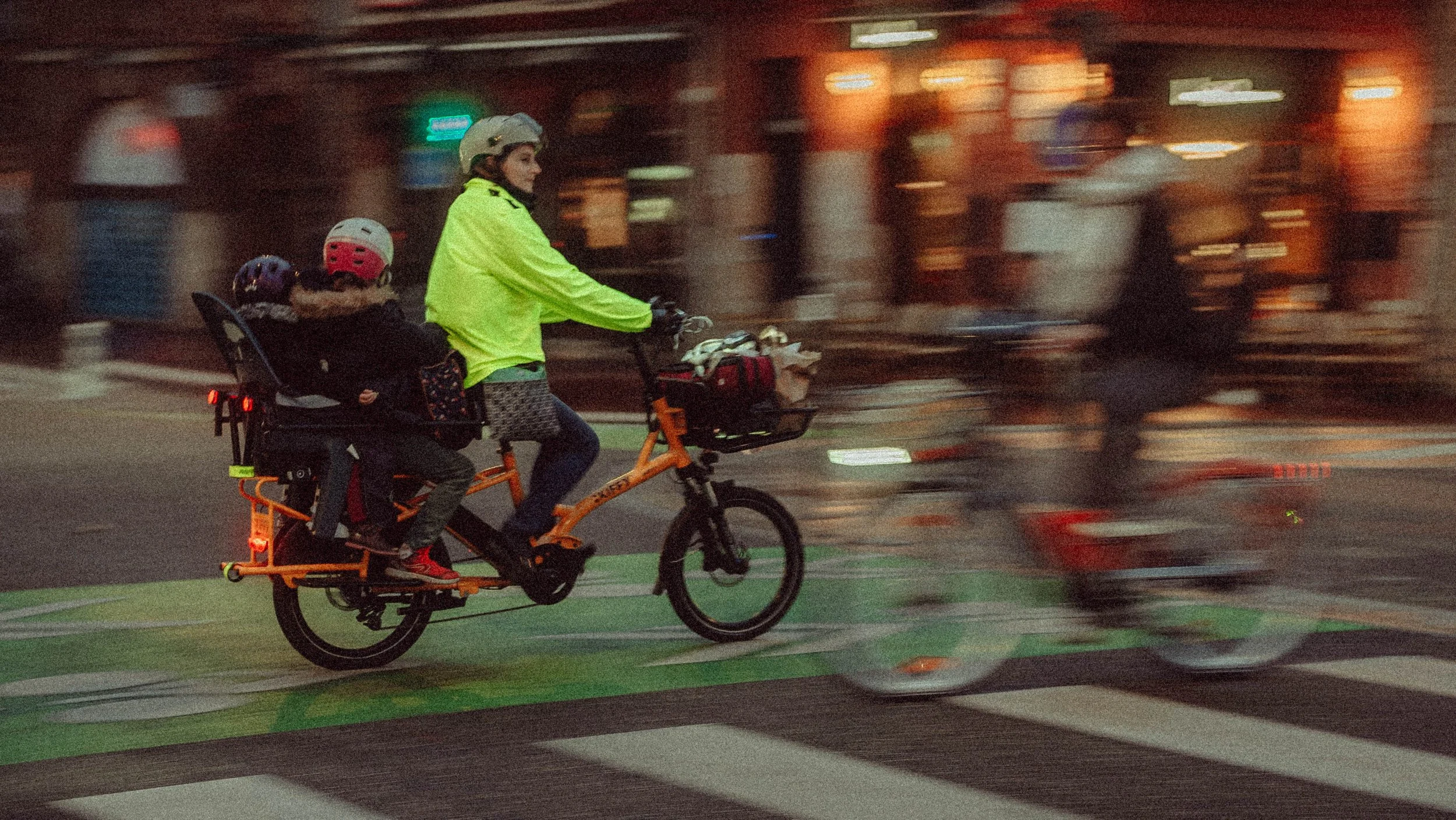 Personne portant des vêtements réfléchissants conduisant un vélo cargo avec deux enfants assis à l'arrière, traversant une rue animée.