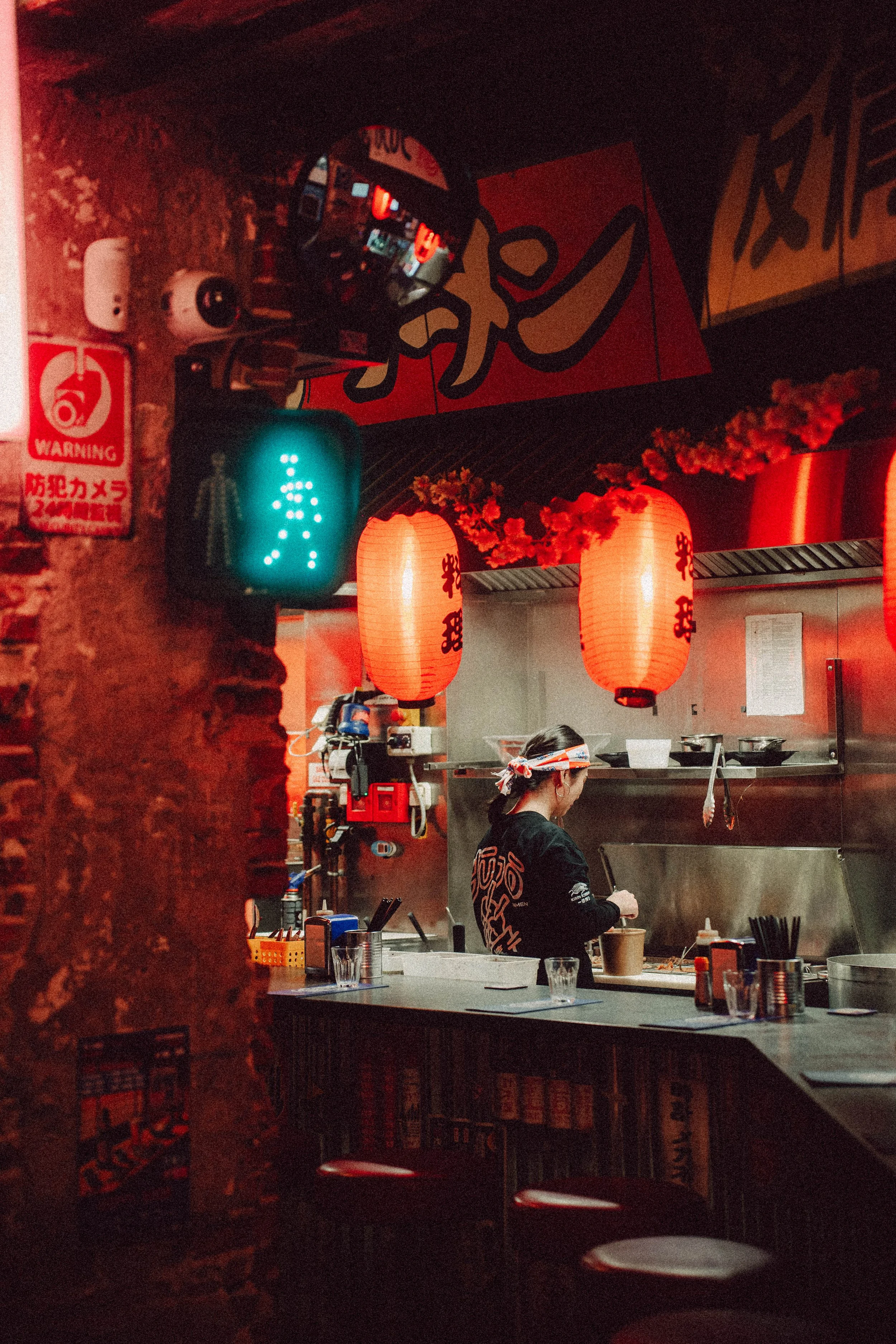Un cuisinier dans un restaurant asiatique avec des lanternes rouges, préparant de la nourriture derrière un comptoir. Une lumière de signalisation verte est visible à gauche de l'image.