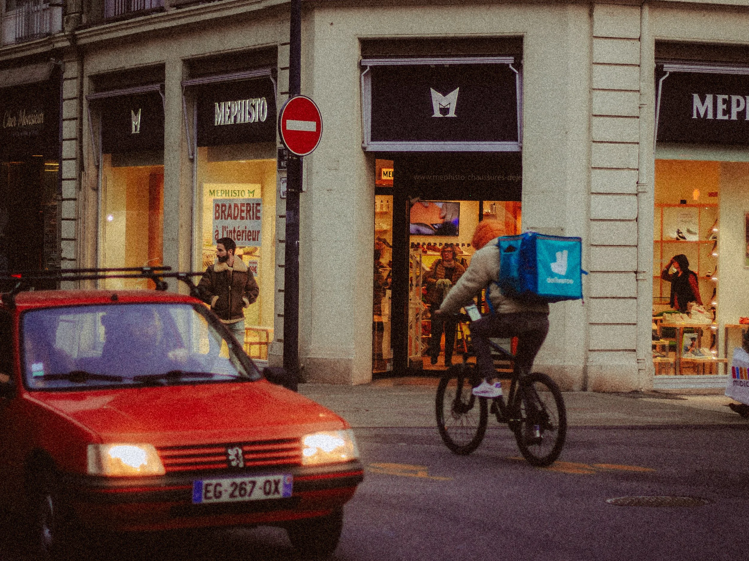 Ruelle animée avec un livreur à vélo portant un sac "Deliveroo", une voiture rouge passant, et des boutiques avec des vitrines éclairées, dont une affiche de "braderie" visible à l'entrée.