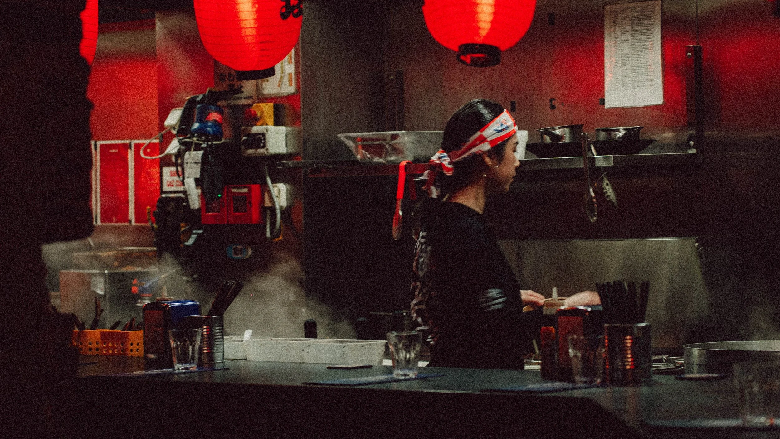 Une cuisinière asiatique dans un restaurant, portant un bandana rouge et blanc, derrière le comptoir de cuisine. Des lanternes rouges pendent au plafond.