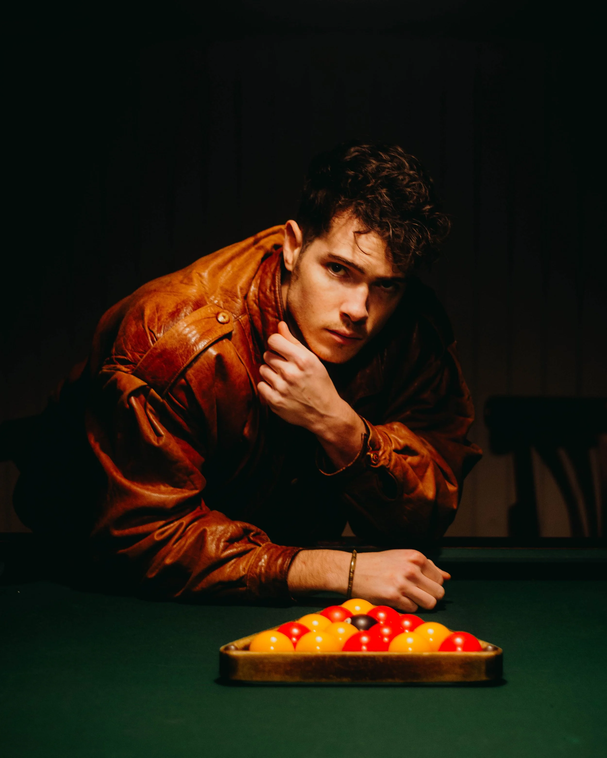 Un homme en blazer en train de jouer au billard, avec une table de billard et un plateau de billes colorées devant lui, dans une pièce sombre.