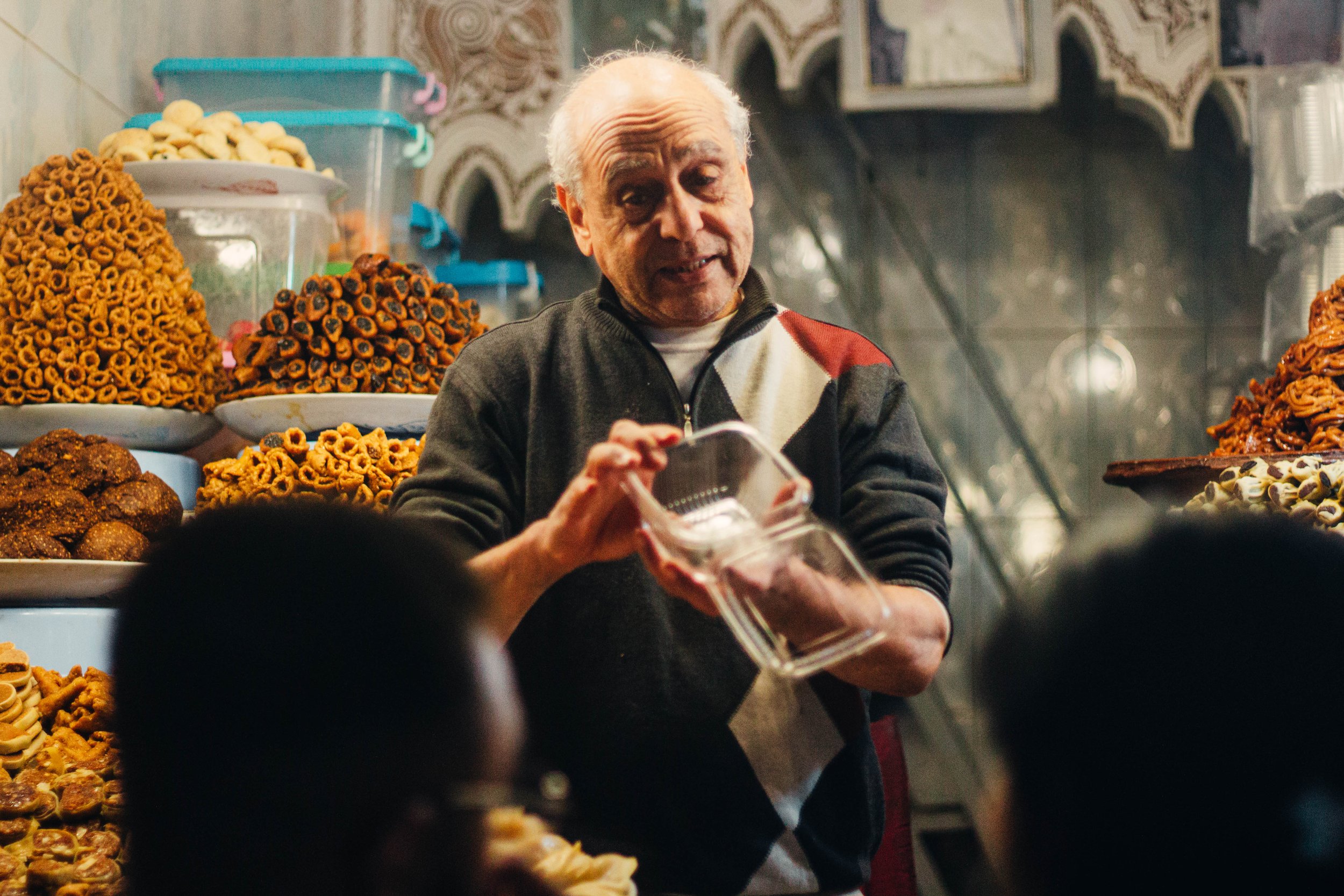 Un vendeur âgé dans une boutique de pâtisseries traditionnelles, présentant des gâteaux variés sur des assiettes empilées, avec des décorations en arrière-plan.