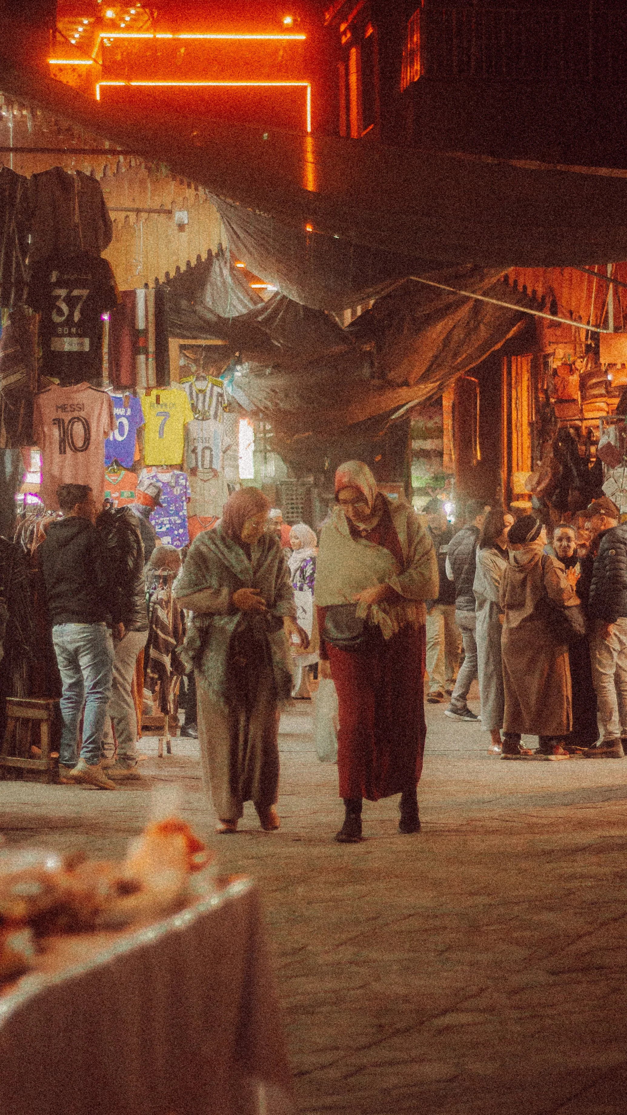 Scène de marché de nuit avec des gens marchant, des vêtements suspendus, ambiance chaleureuse.