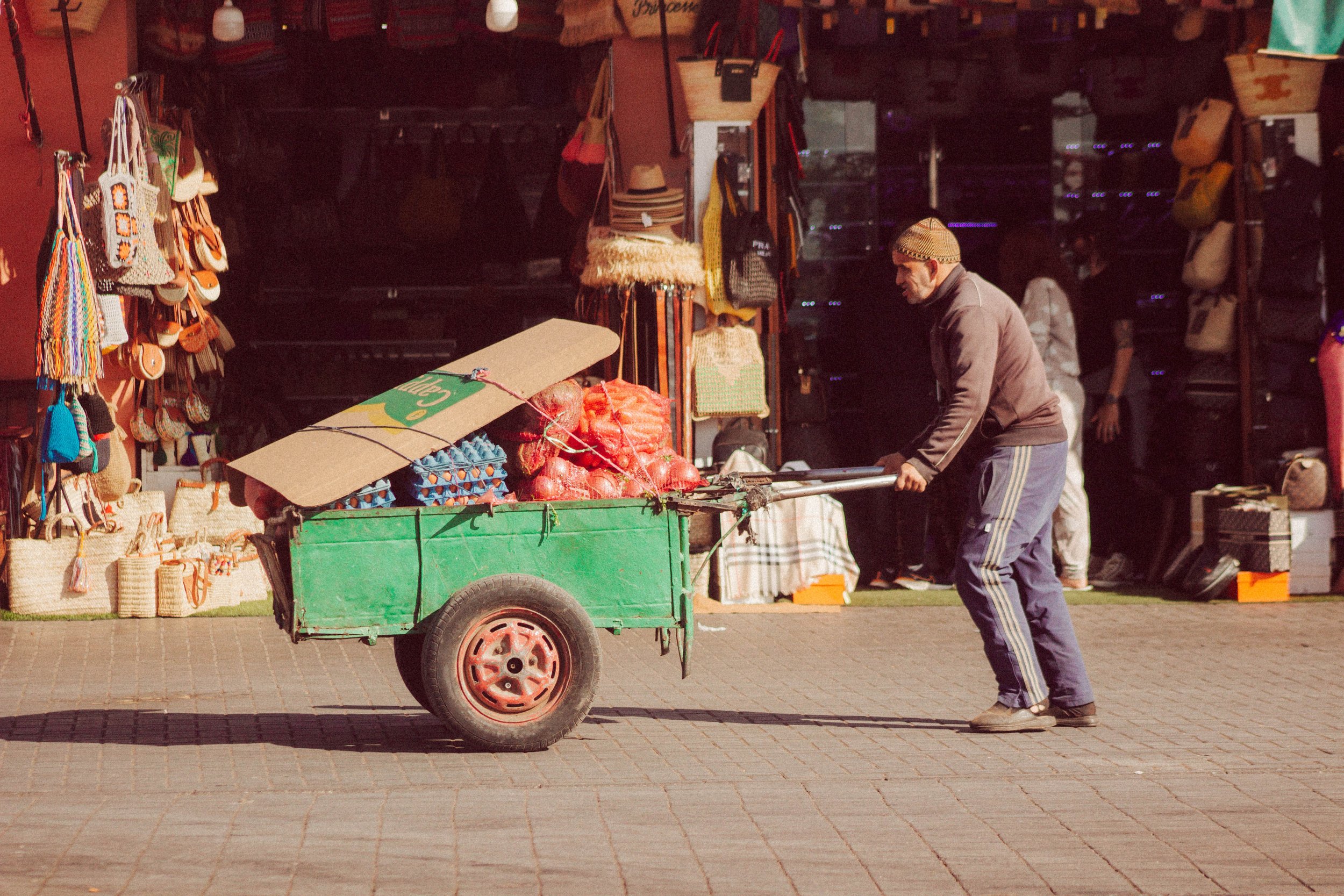 Homme poussant un chariot chargé de marchandises, devant des boutiques de sacs et accessoires.