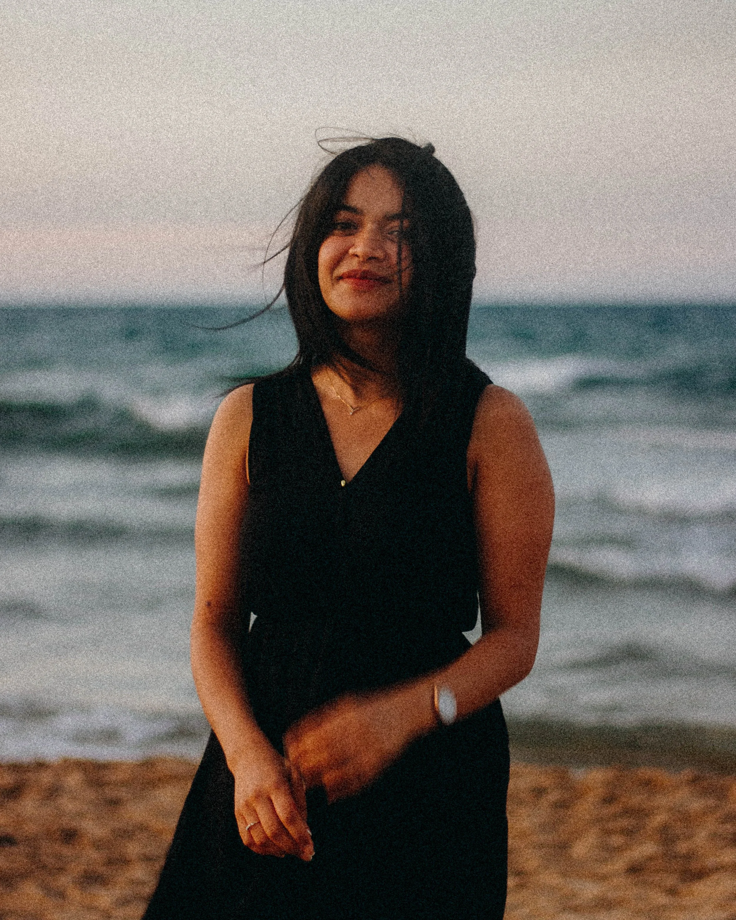 Femme souriante en robe noire sur une plage au bord de la mer.