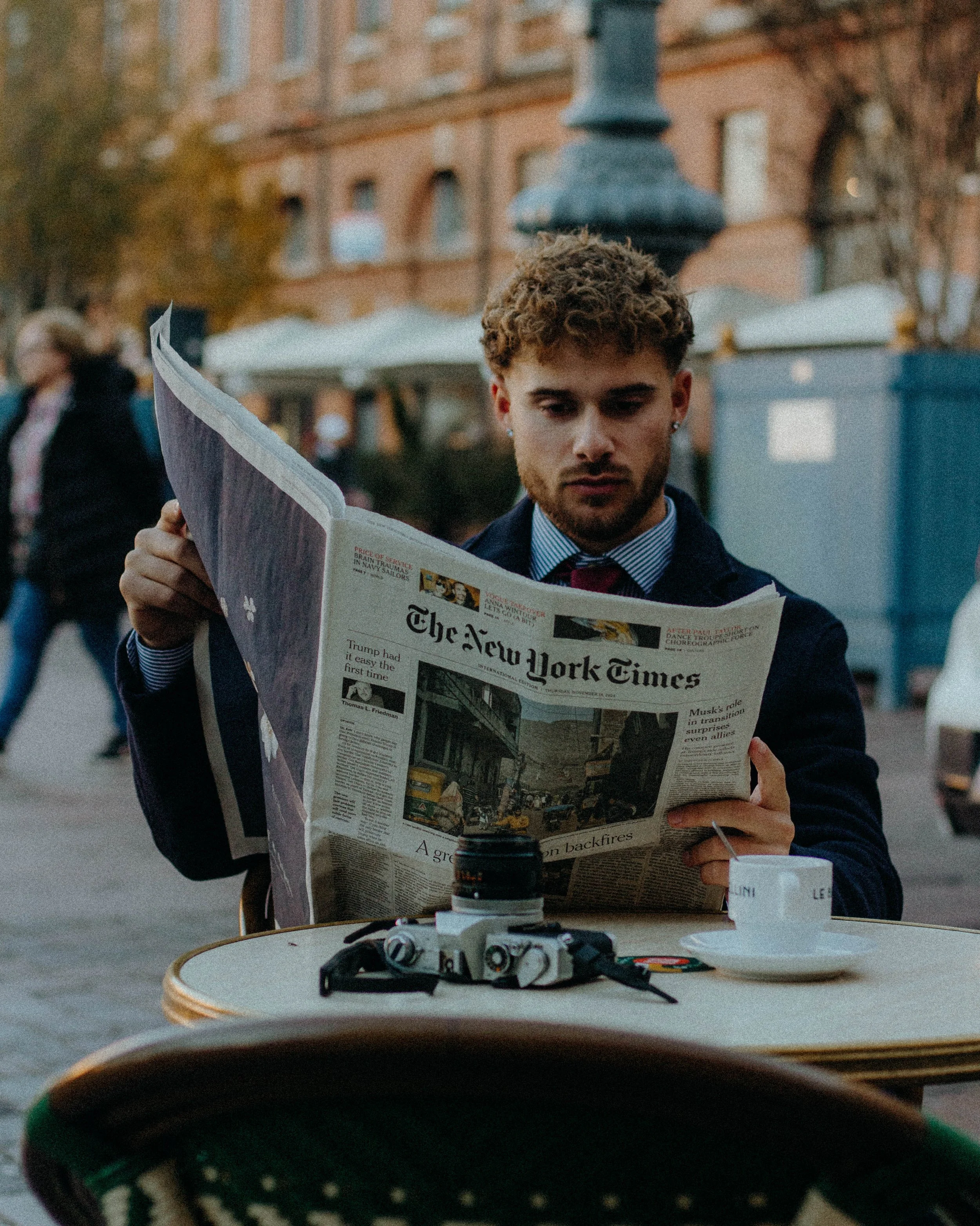 Homme lisant un journal à une table de café, avec un appareil photo et une tasse de café posés devant lui, en extérieur urbain.
