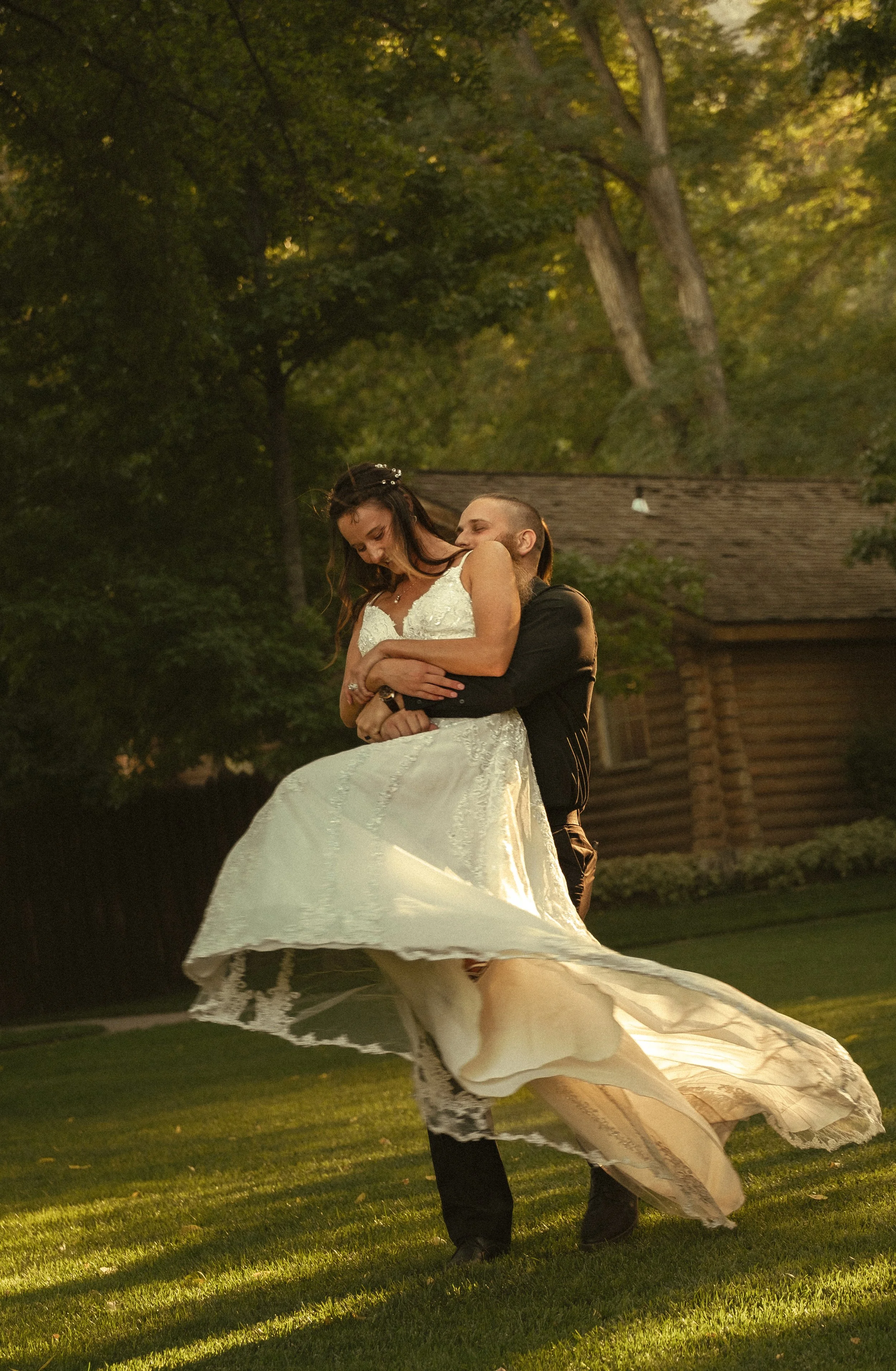 Man in black shirt lifting woman in white wedding dress on grassy field with trees and log cabin in background.