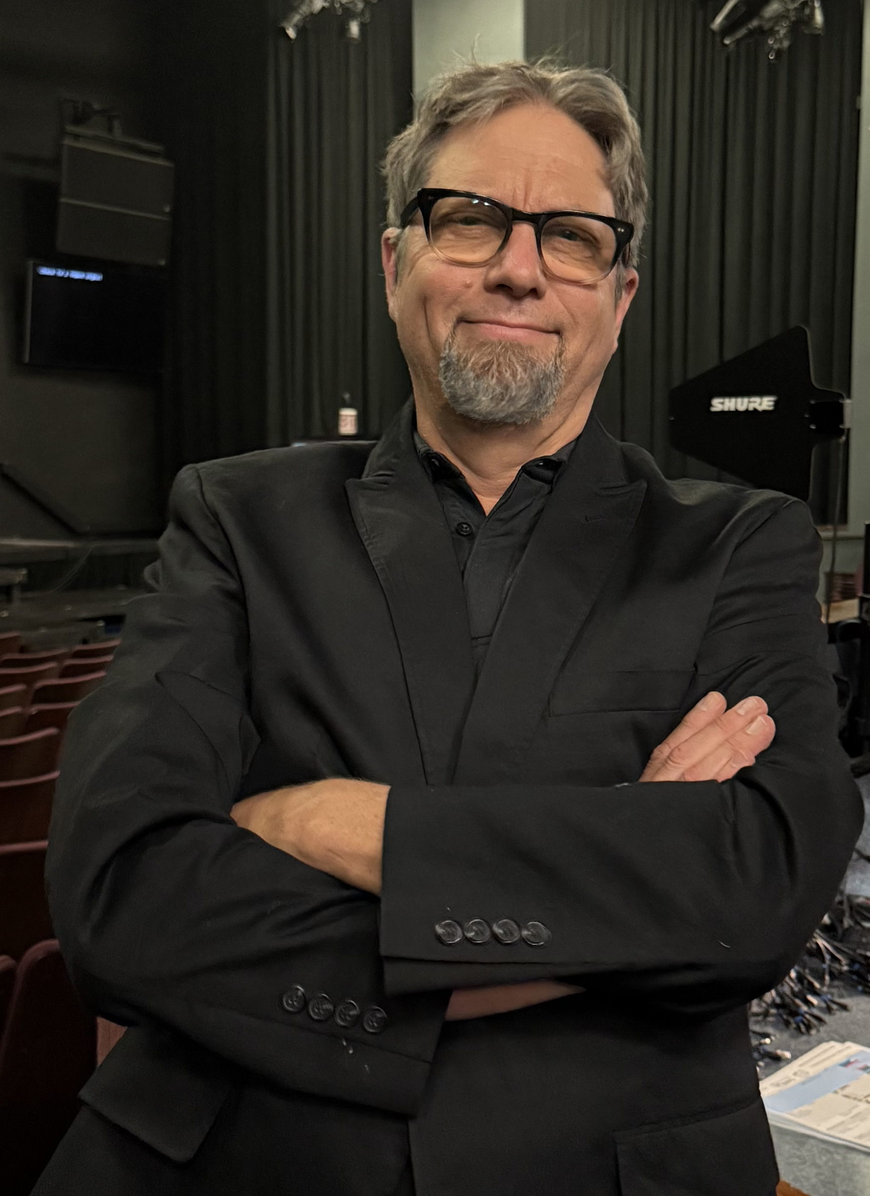Man in black shirt with glasses at sound mixing console, holding a pen and paper, wearing a headset, with monitors and equipment in background.