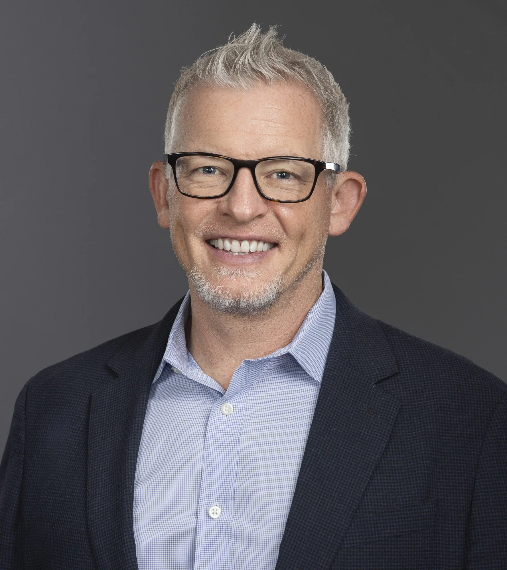 A middle-aged man with short gray hair and glasses, wearing a dark blazer over a light blue shirt, smiling against a gray background.