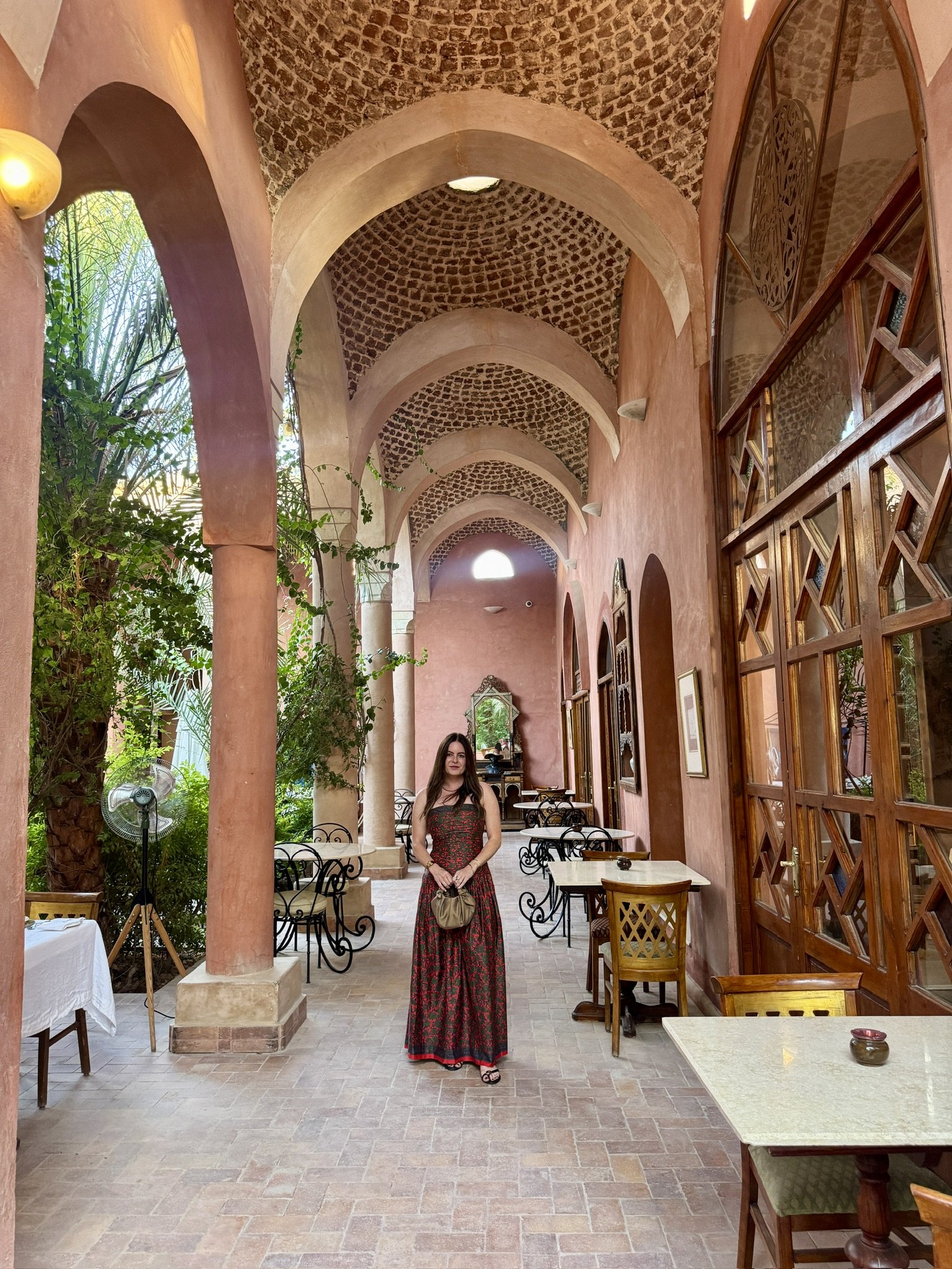 A woman in a red and black patterned dress standing in a Mediterranean-style corridor with arched ceilings, peach-colored walls, and wooden accents. There are tables and chairs along the corridor, with greenery visible on the left side.