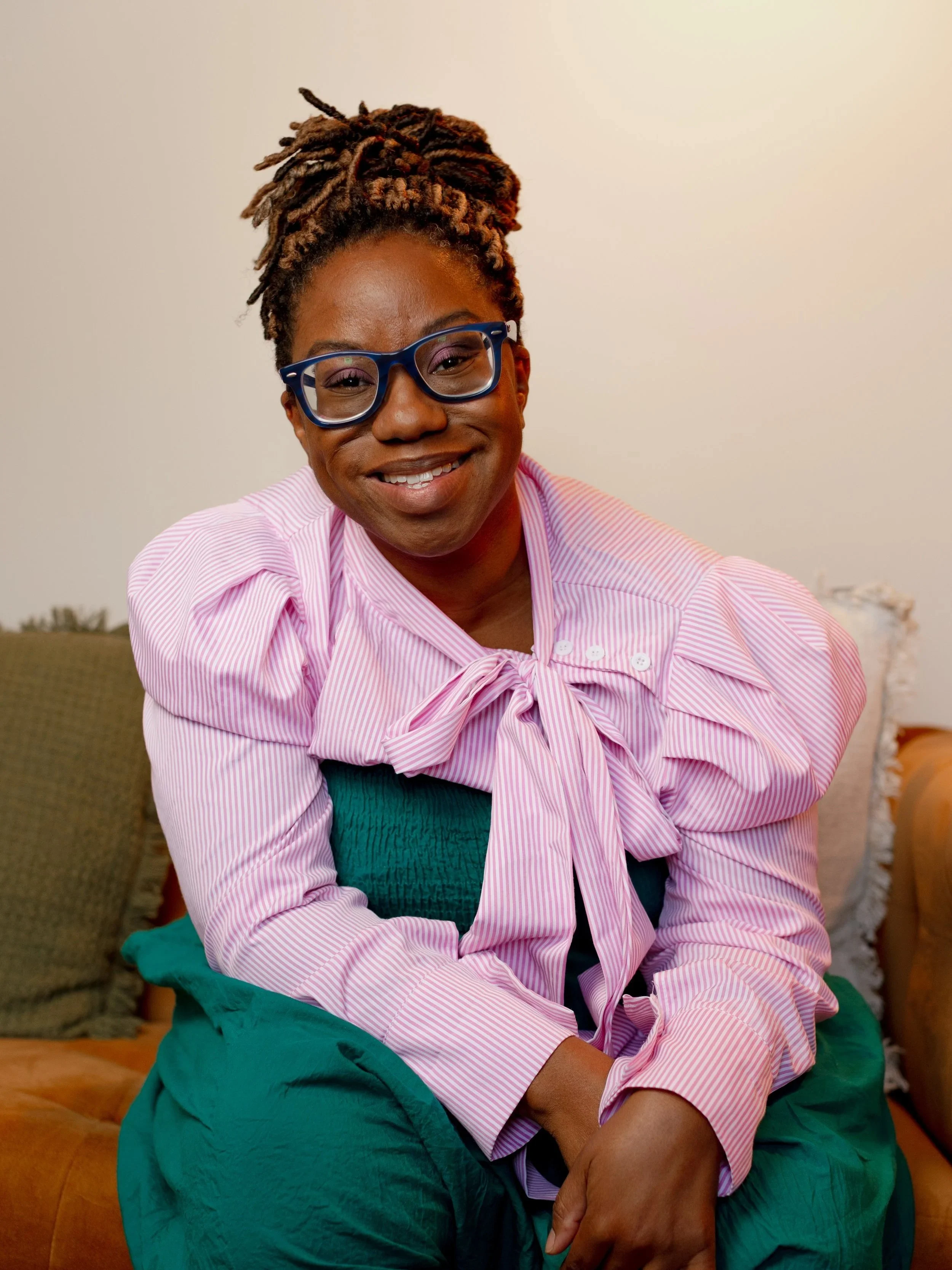 A woman with glasses and styled dreadlocks smiling while sitting on a sofa in a room with beige walls at a psychology clinic.