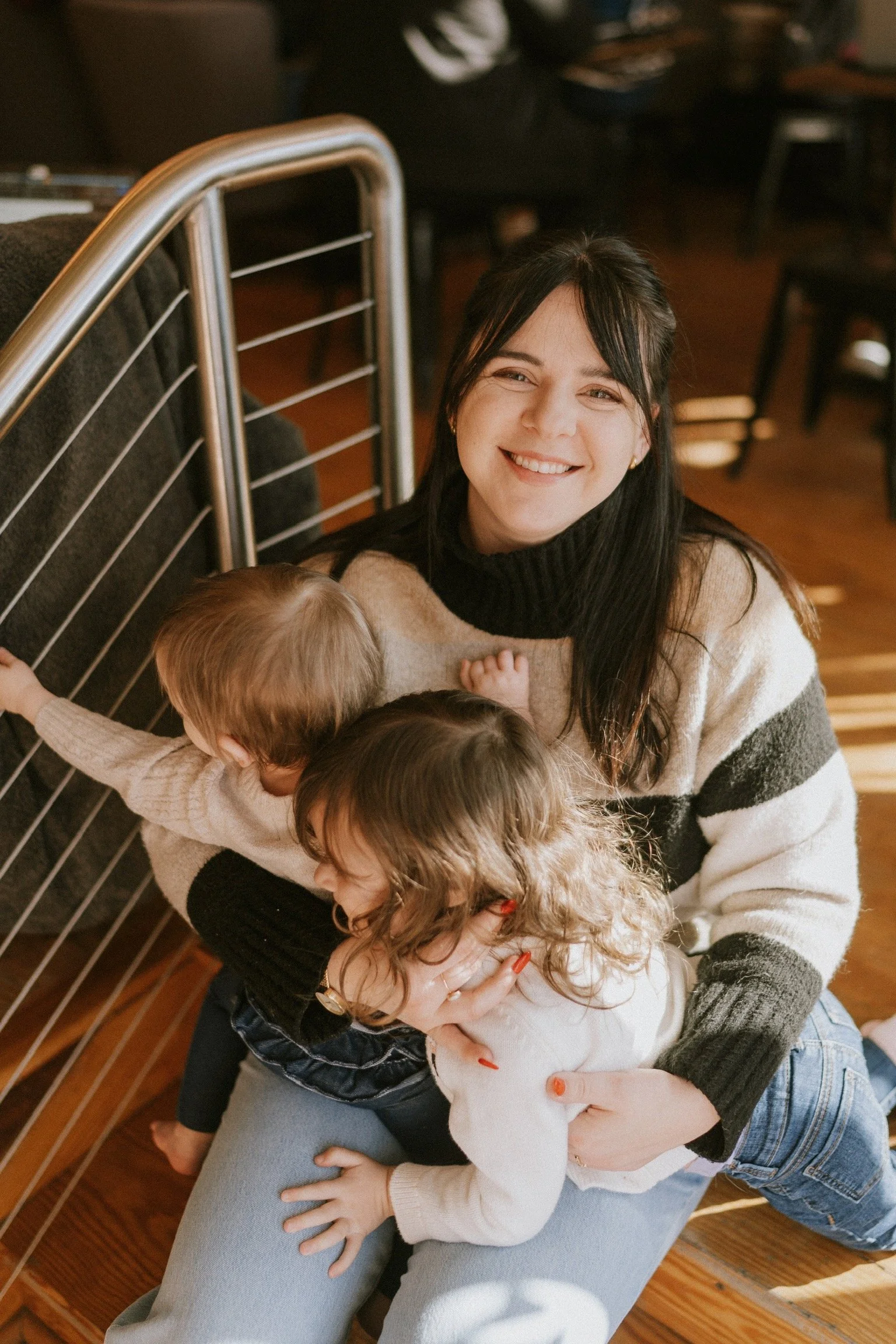 A woman smiling happily while sitting on the floor, holding and hugging three young children, in a cozy indoor setting.