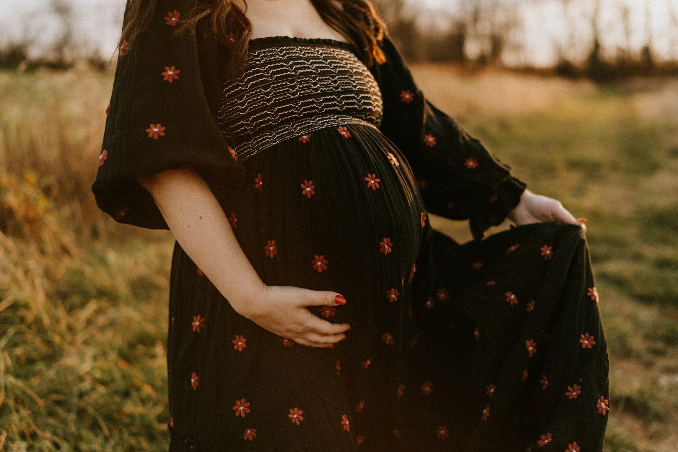 A pregnant woman outdoors in a field holding her belly with one hand and lifting her dress with the other. She wears a black dress with red floral patterns and the background is a natural setting with trees and grass during sunset.
