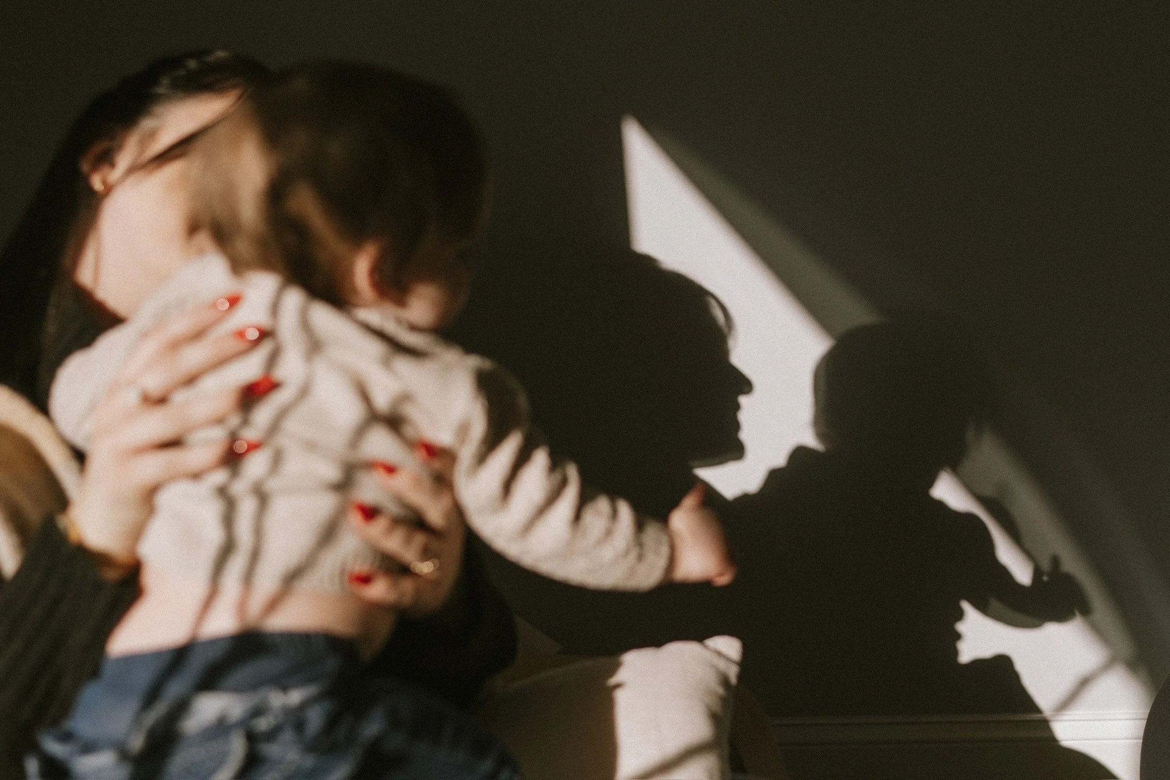 Woman holding child with sunlit shadows creating silhouettes on the wall.