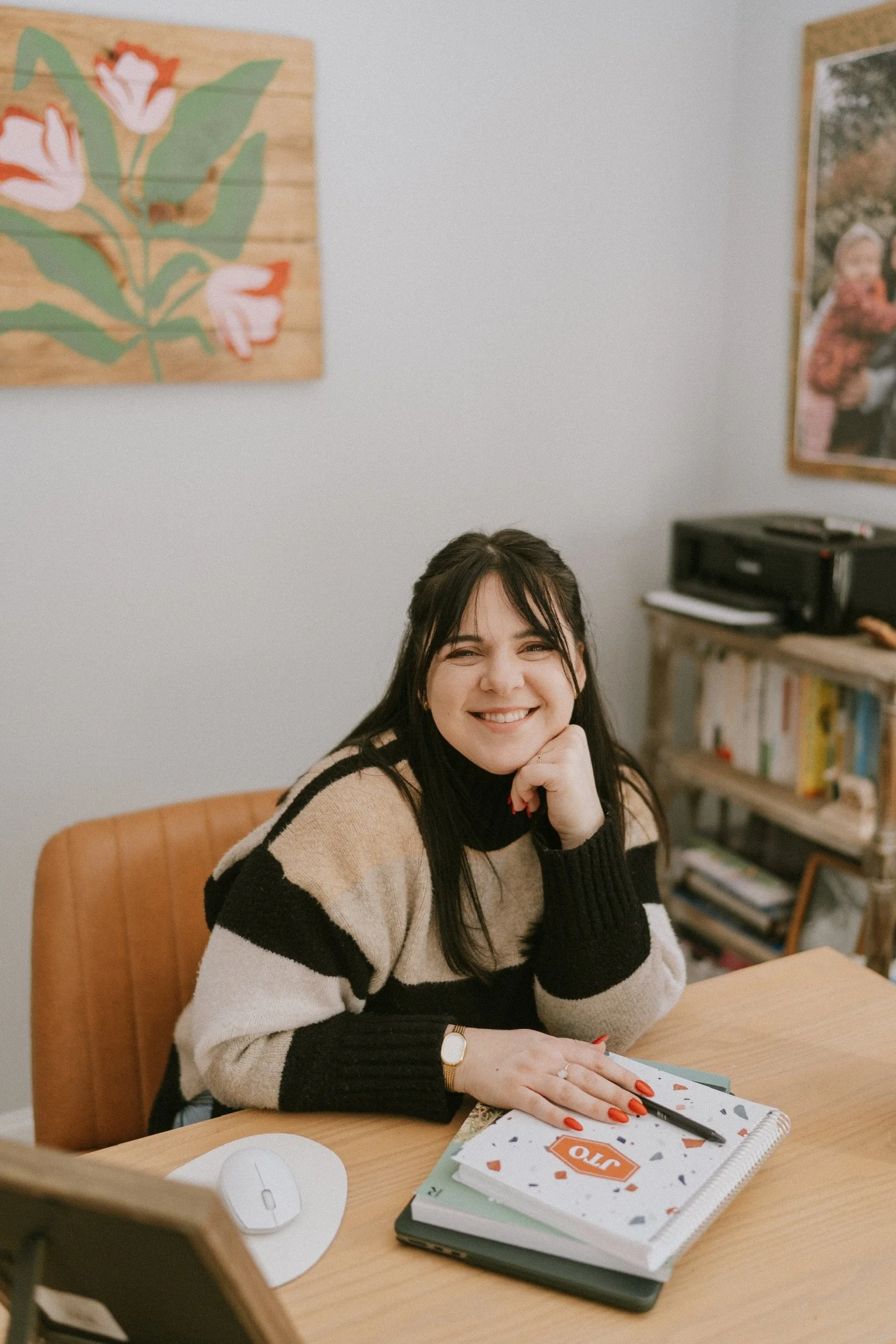 A woman with dark hair smiling while sitting at a wooden desk with a notebook, a pen, and a computer mouse, in an office or home workspace.