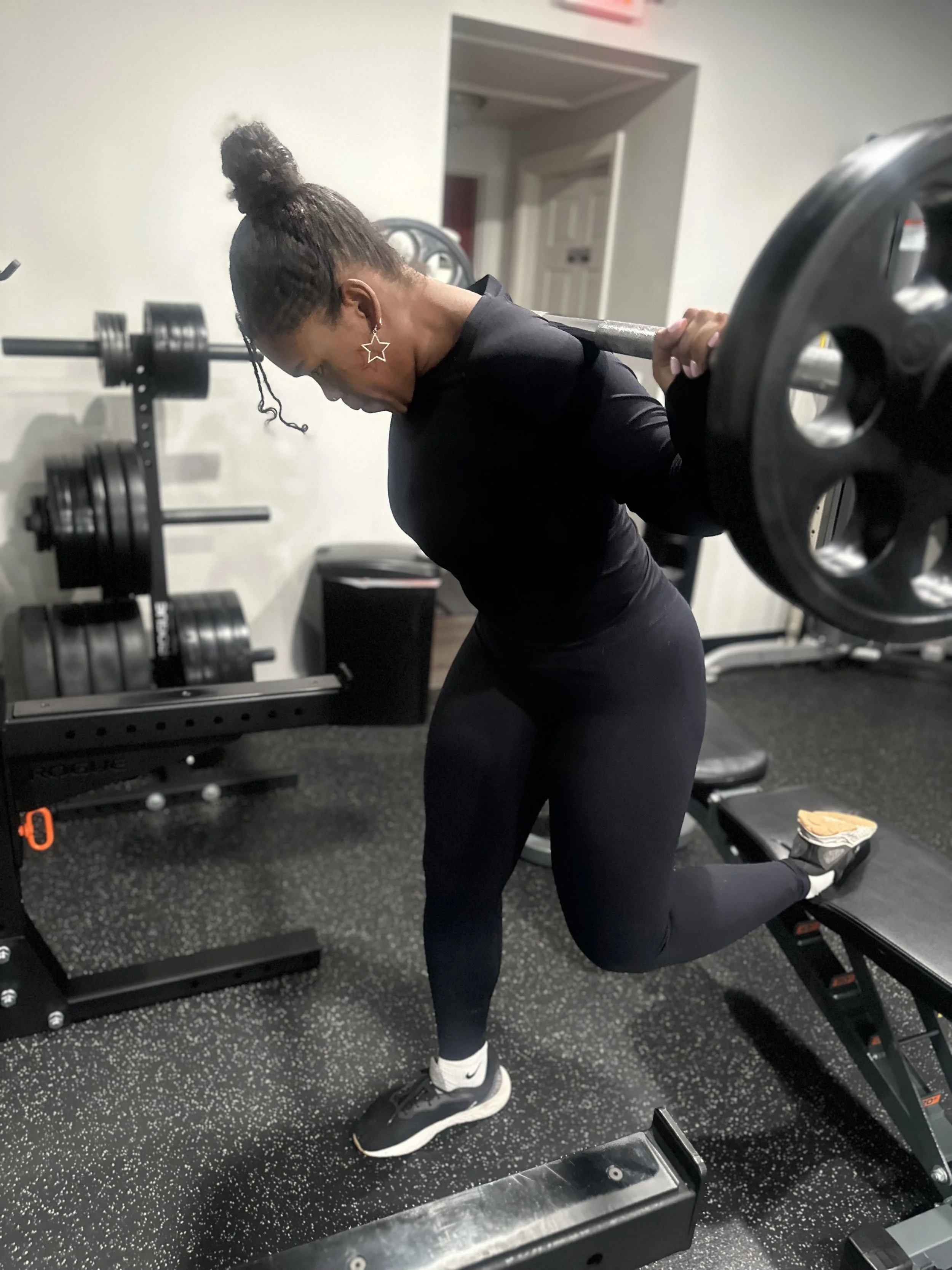 A woman with dark hair in a bun and star-shaped earrings is performing a barbell lunge exercise in a gym, wearing black workout clothes and sneakers, with gym equipment and weights in the background.