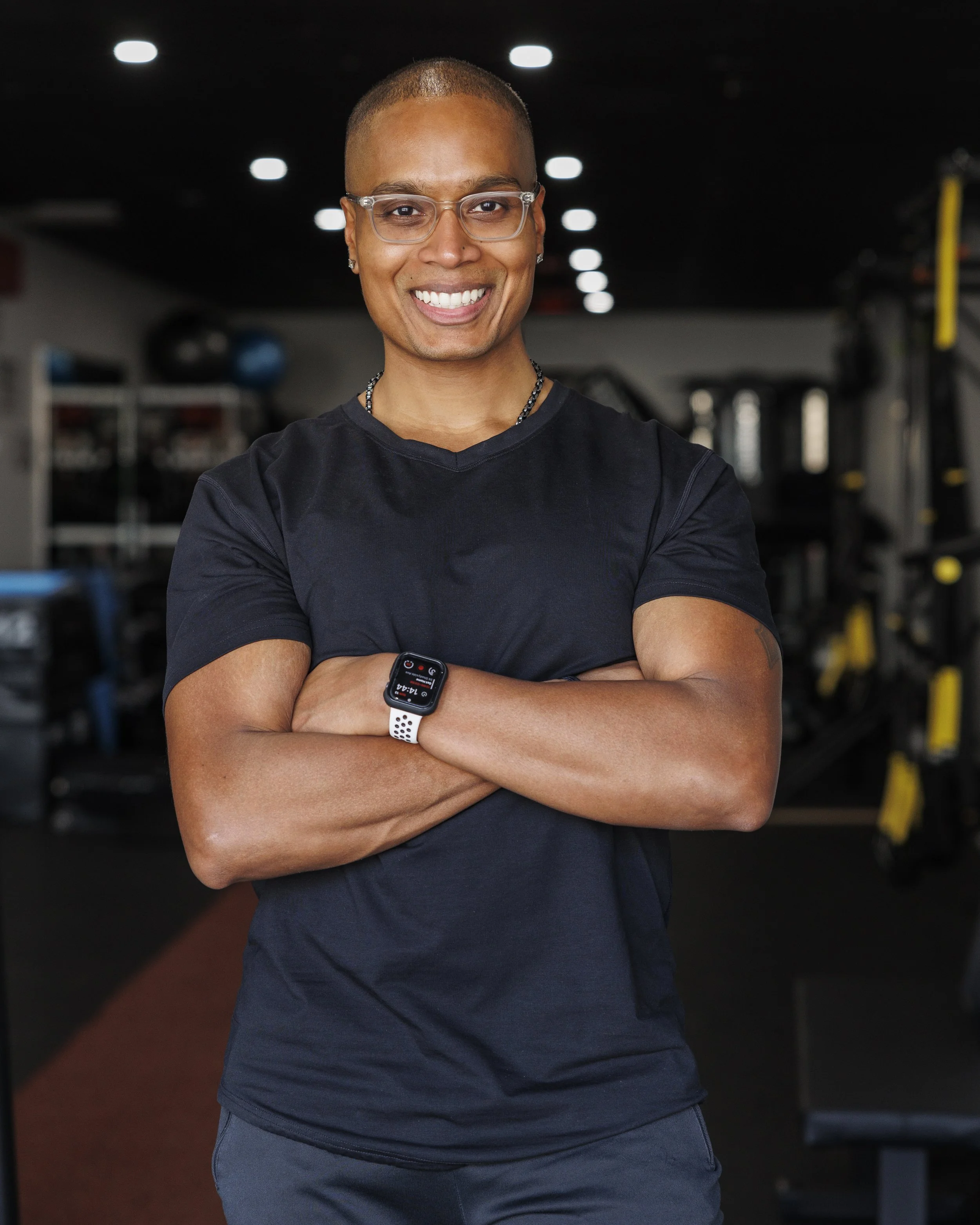 Smiling person standing with arms crossed in a gym, wearing glasses, a black shirt, a smartwatch, and jewelry.