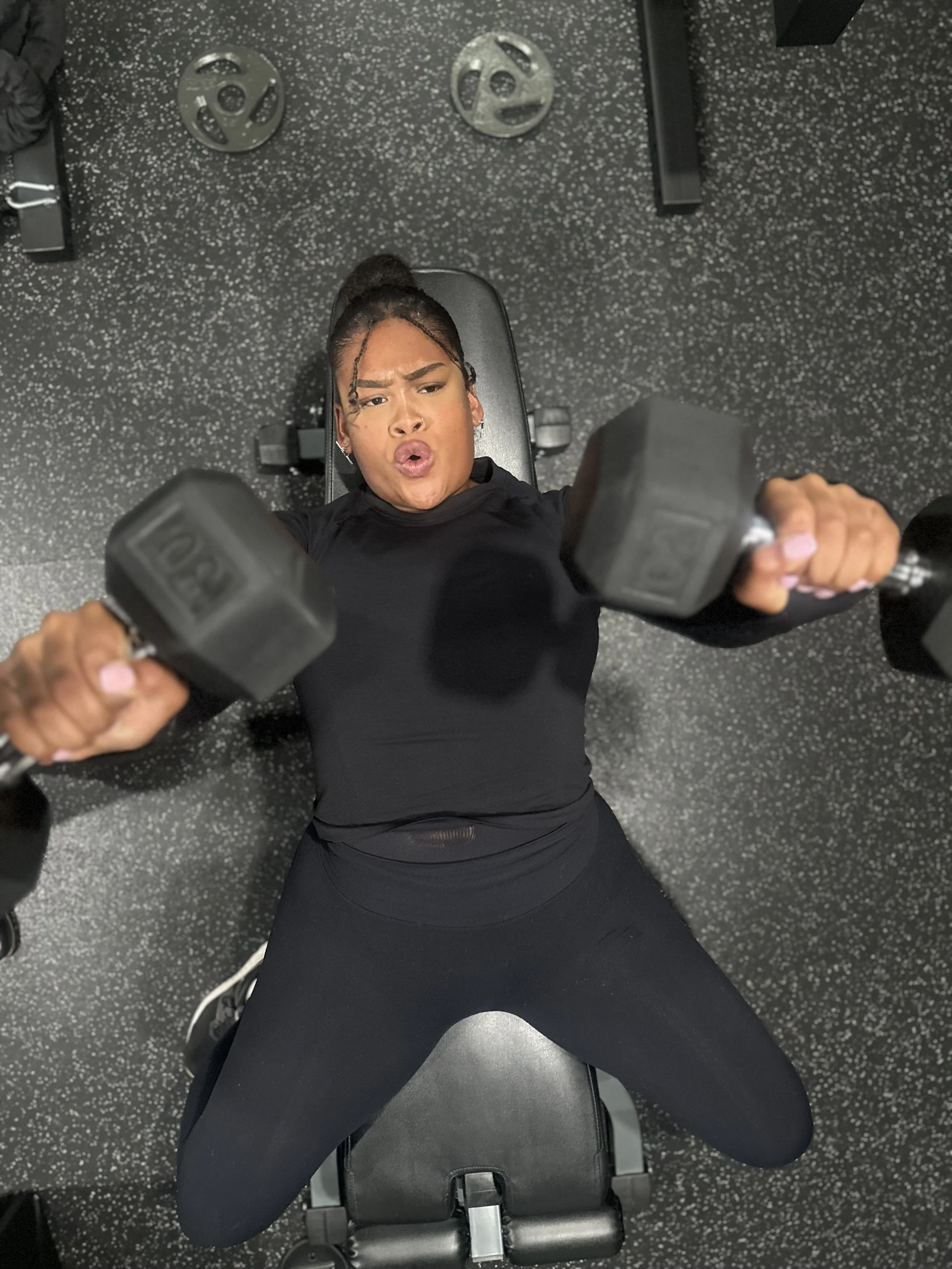 Woman in black workout clothes lying on a bench lifting dumbbells in a gym.