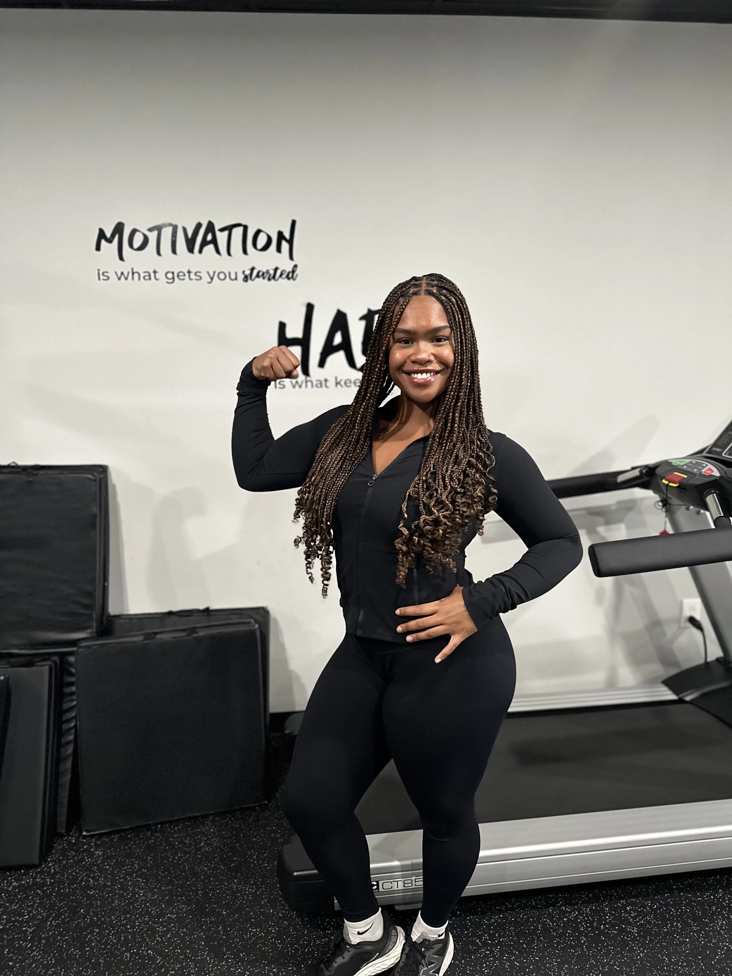 A woman in black workout clothes flexing her arm in a gym with motivational quotes on the wall behind her.