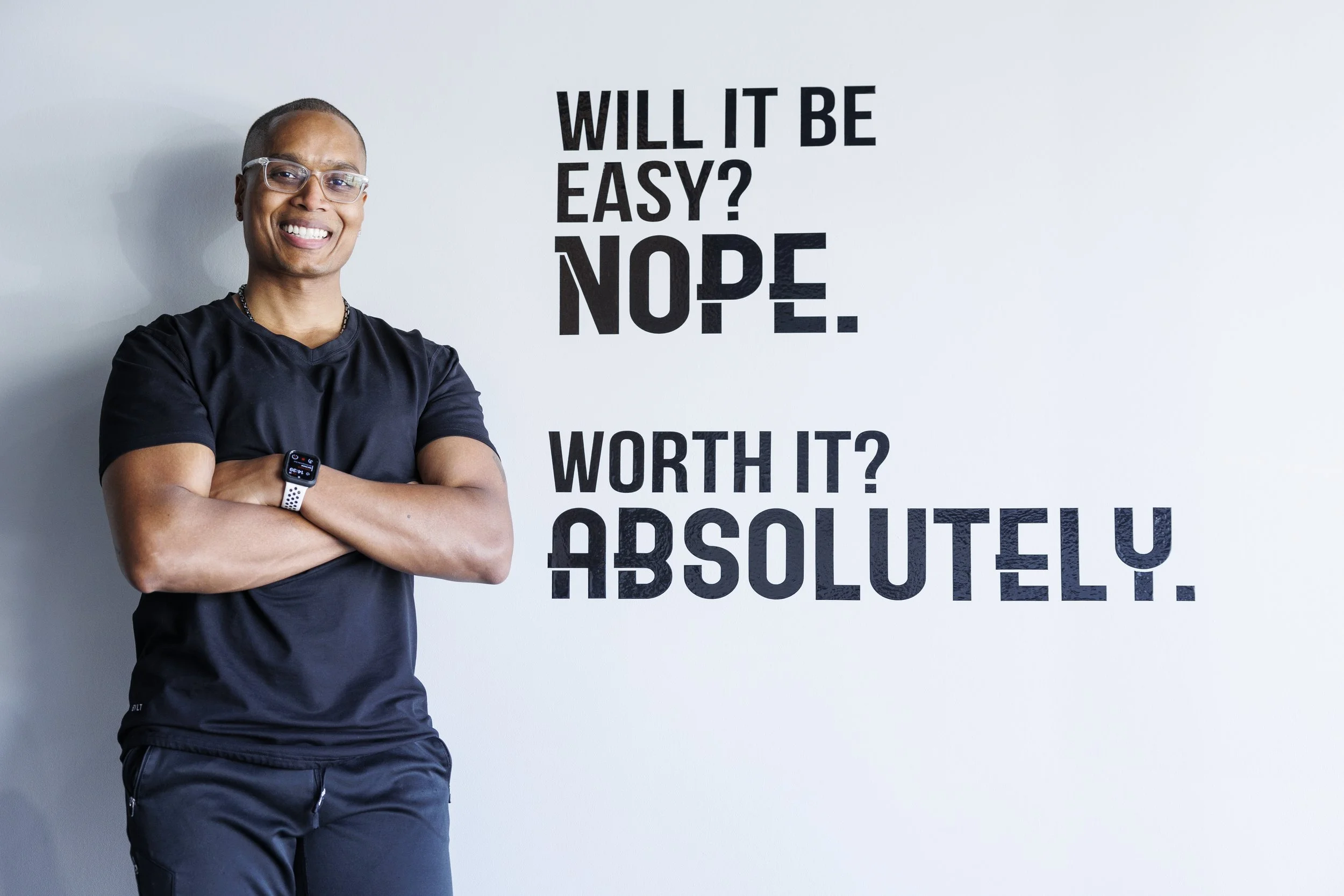 A smiling man with glasses, wearing a black T-shirt and smartwatch, standing with crossed arms beside a white wall with text that reads, "Will it be easy? Nope. Worth it? Absolutely."