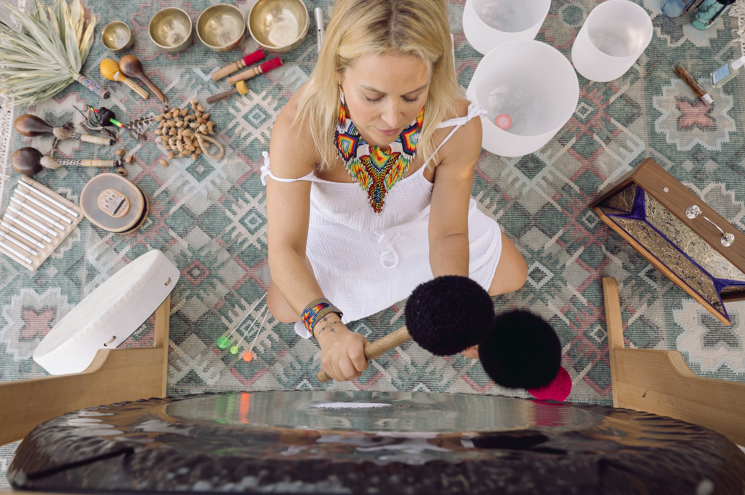 A woman with blonde hair wearing a white dress and colorful jewelry, sitting on the floor with musical instruments and singing bowls around her, holding a mallet, possibly performing a sound healing session.