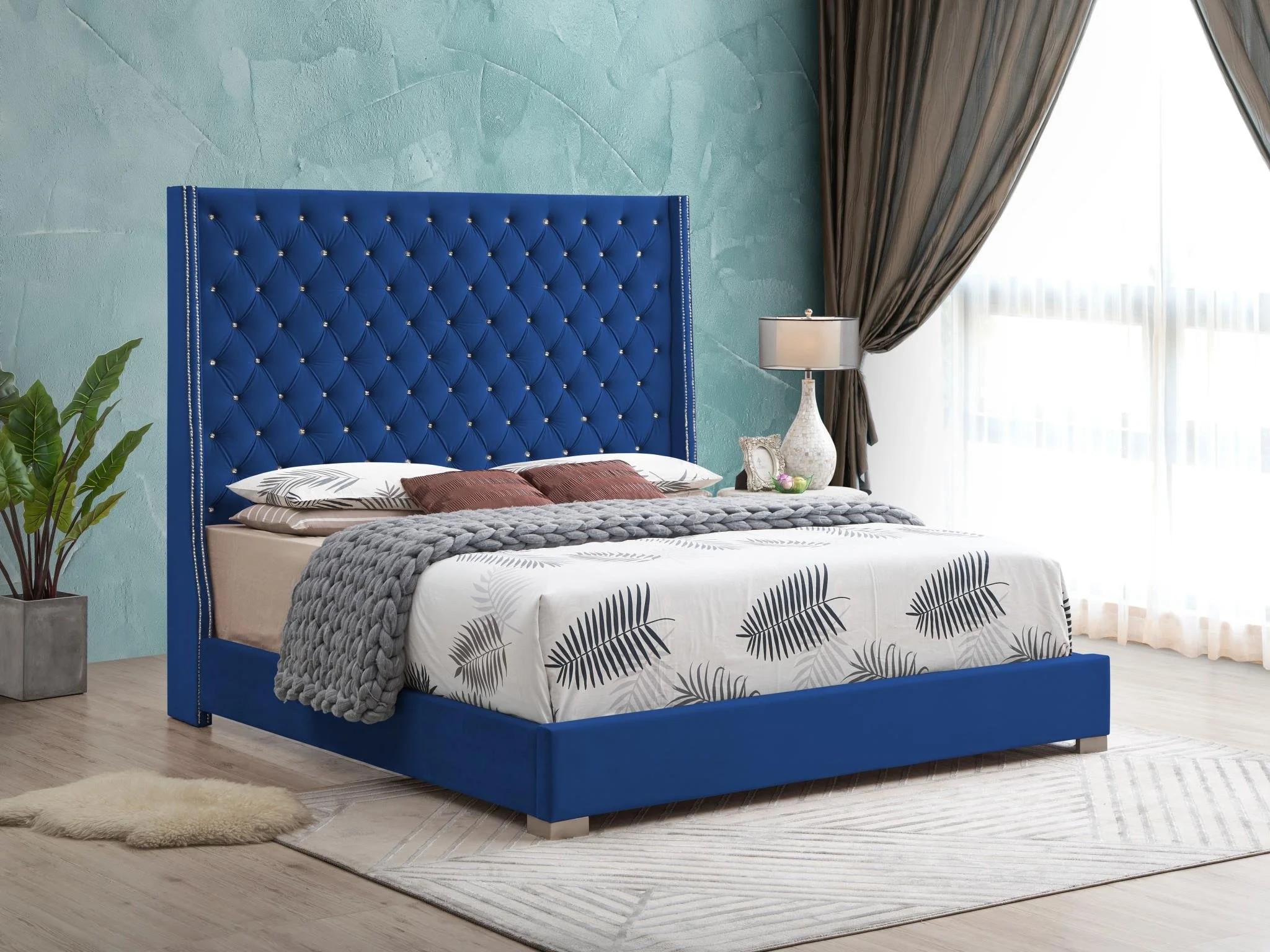 A bedroom with a large blue upholstered bed featuring a tufted headboard. The bed has white and gray botanical patterned bedding, brown pillows, and a chunky gray throw blanket. To the right of the bed is a white table lamp with a beige shade, a small framed picture, and a decorative bowl. A sheer curtain and darker drapes cover a window behind the lamp, with a potted plant on the left side of the room. The floor is light wood with a cream-colored rug and a small faux fur rug near the bed.