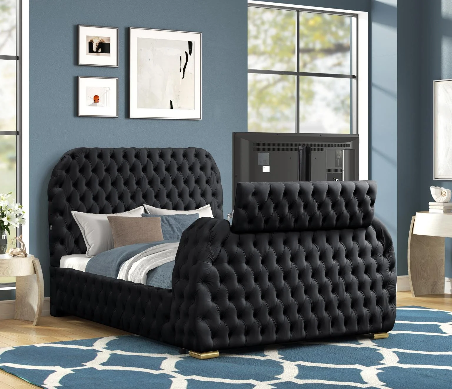 A bedroom with a black, tufted, upholstered bed against a blue wall. The bed has white, beige, and navy pillows, and is covered with a light blue blanket. There is a large window with sunlight illuminating the room. On the wall above the bed, there are four framed pictures. A black TV is positioned at the head of the bed, facing upwards. To the left, there is a small round side table with a white vase of white flowers. To the right, a light-colored nightstand has a decorative item and a stack of books. A blue and white patterned area rug is underneath the bed.