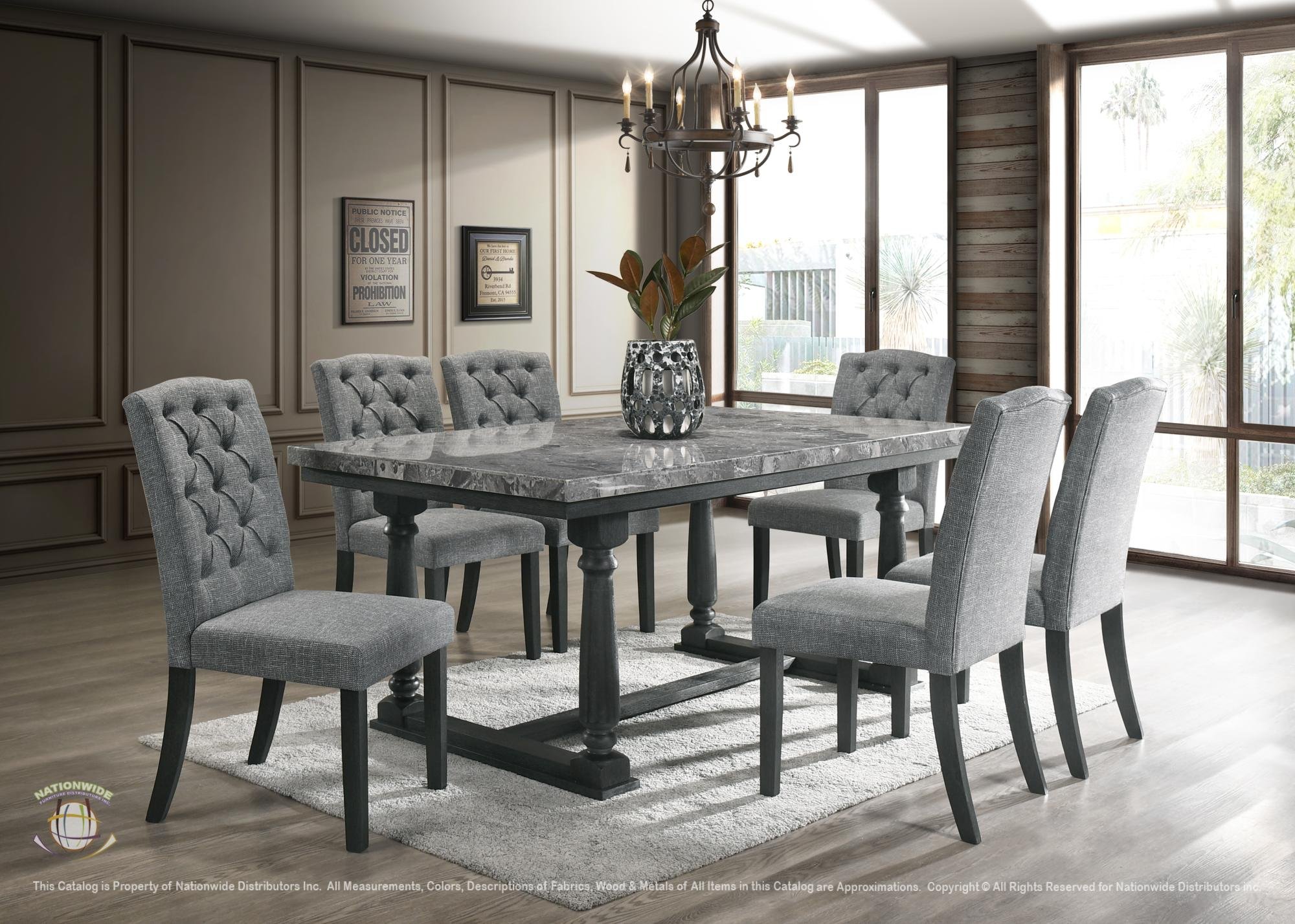 A modern dining room with a rectangular marble-top table surrounded by eight grey upholstered chairs, a black chandelier hanging above, a potted plant centerpiece, and large windows allowing natural light, with wood-paneled walls and framed artwork.