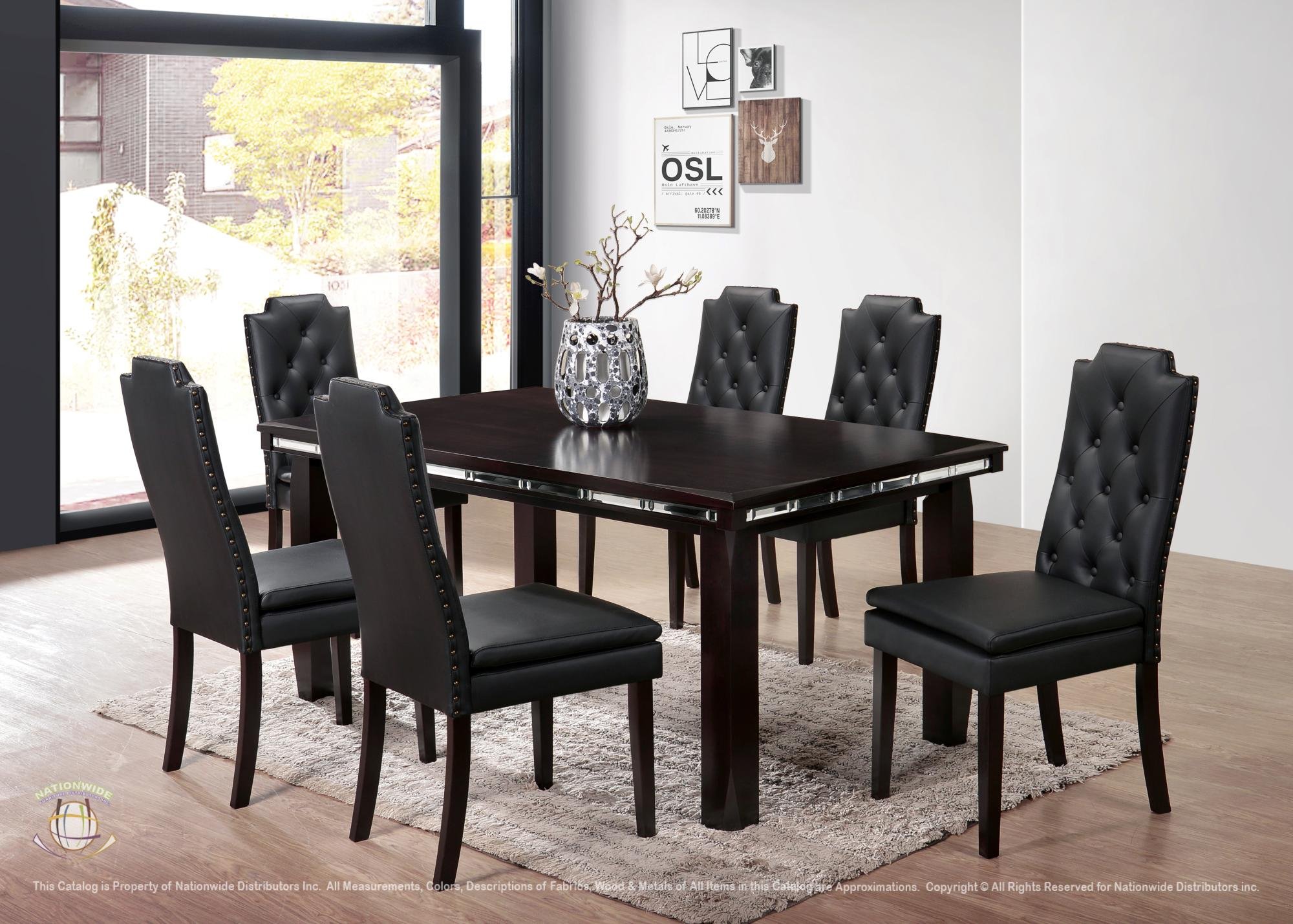 A modern dining room with a dark wood table surrounded by six black leather chairs with button tufting. A decorative vase with branches sits on the table. There are framed artworks on the white wall and large glass doors letting in natural light.