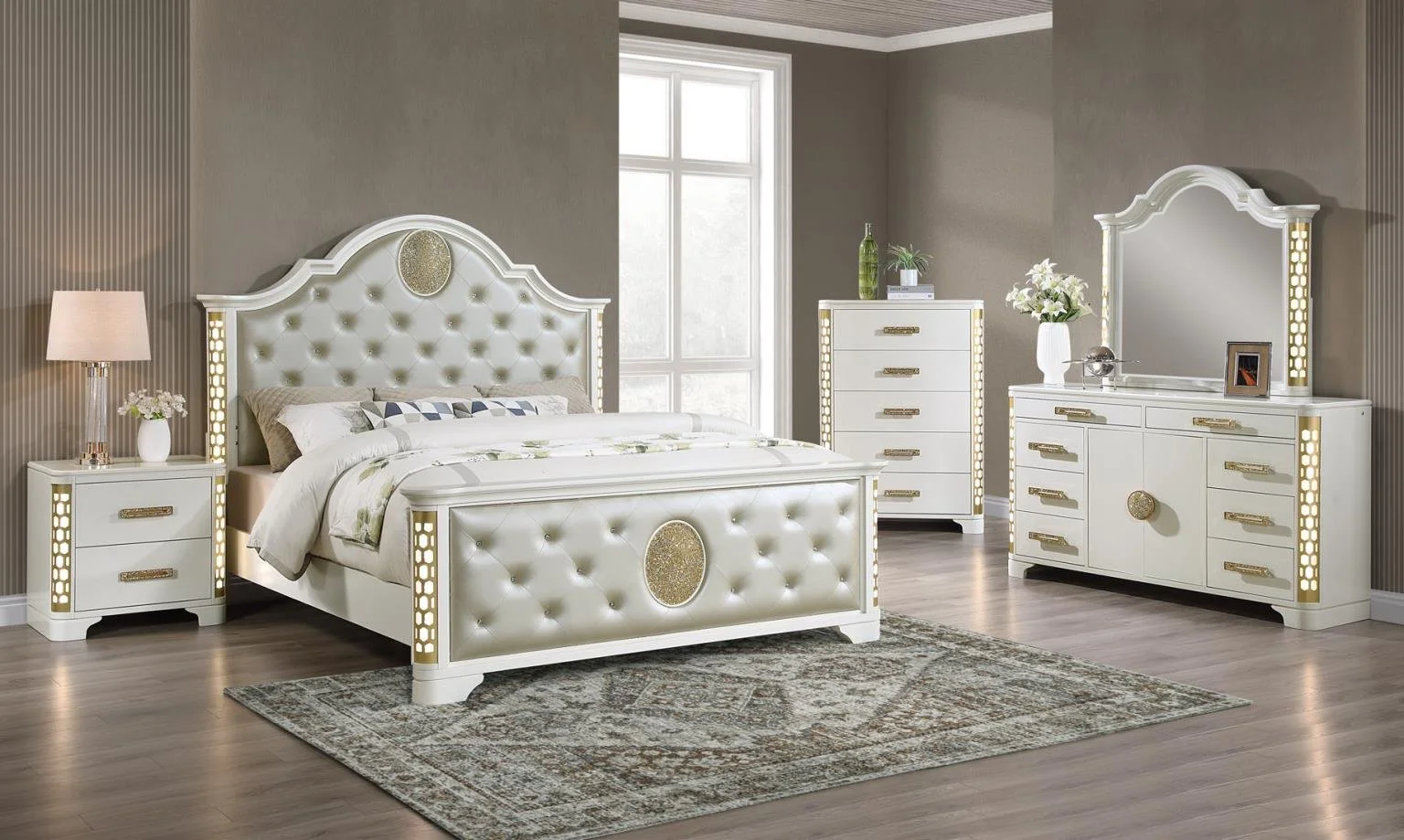 A bedroom with a white, tufted bed with gold accents, a matching nightstand with a lamp and flowers, a tall dresser with a potted plant on top, and a white dresser with a mirror and decorative items, all on a wood floor with a patterned rug.