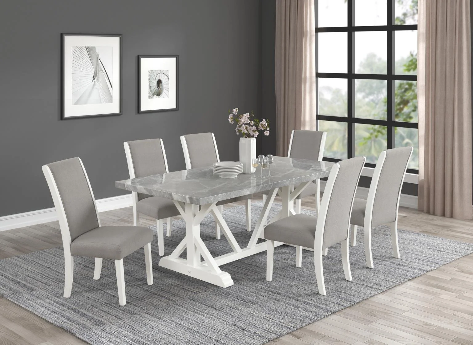 Modern dining room with a marble table, gray upholstered chairs, a gray area rug, and framed black and white artwork on a dark gray wall. Large window with beige curtains shows greenery outside.