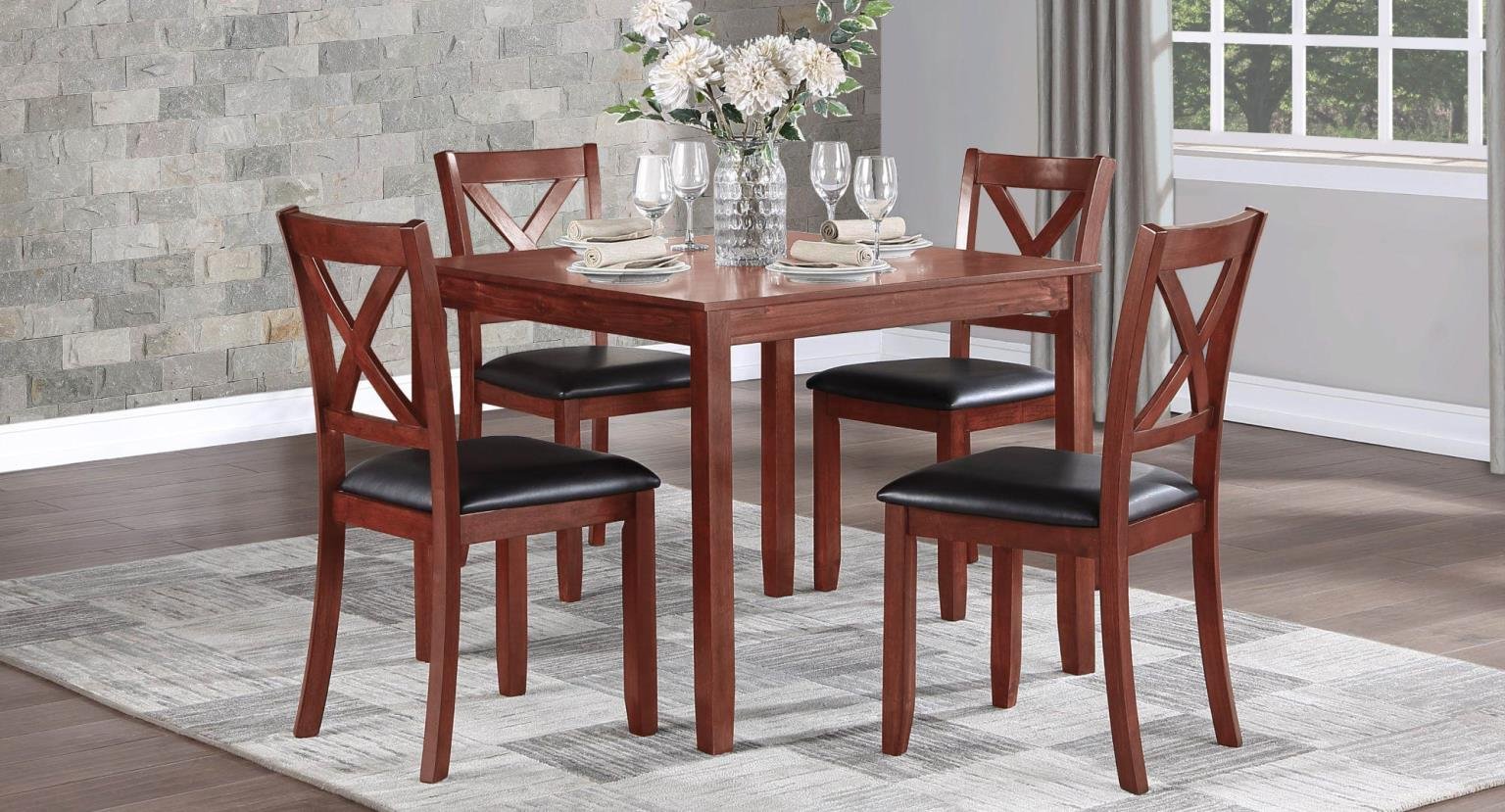 A dining room table set with four chairs, a large vase of white flowers, four wine glasses, napkins, and plates, in a room with wood flooring, a textured gray stone wall, and a window with a view of trees.