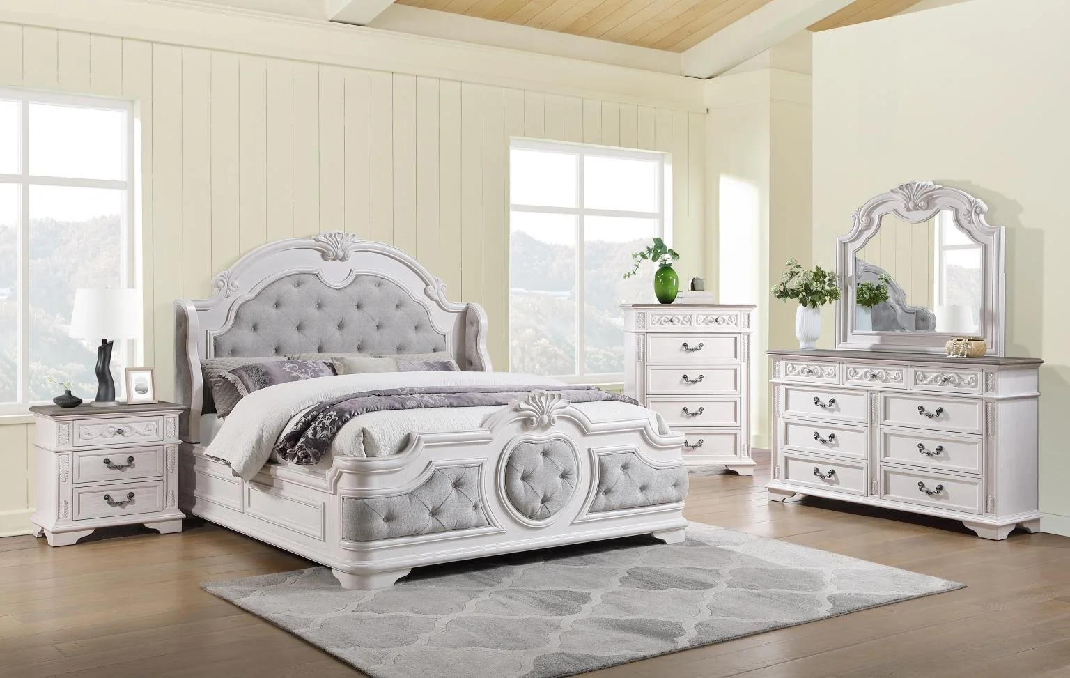 A bedroom with a large white bed that has an ornate headboard and footboard, gray tufted upholstery, and gray bedding. There are two white nightstands with lamps, a white dresser with a mirror, and a white chest of drawers with a green vase and flowers. The room has light yellow walls, wooden flooring, and large windows letting in natural light.