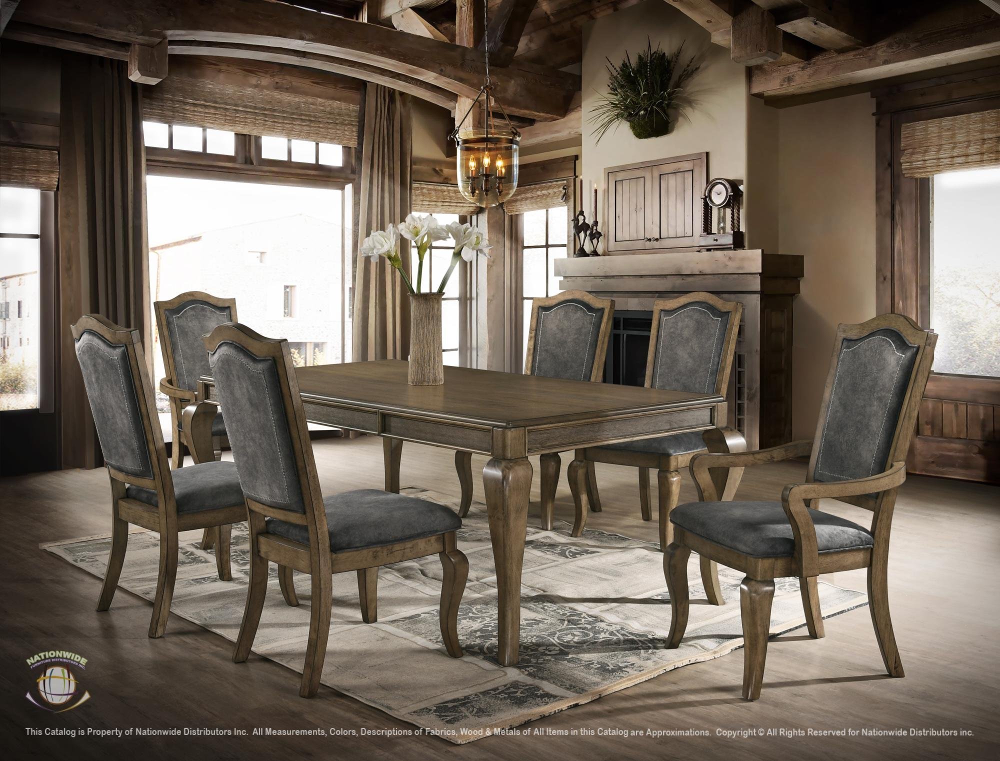 A rustic dining room with six wooden chairs around a rectangular table, a vase with white flowers on the table, large windows with curtains, a fireplace above which is a floating cabinet and decorative items, and wooden beams on the ceiling.
