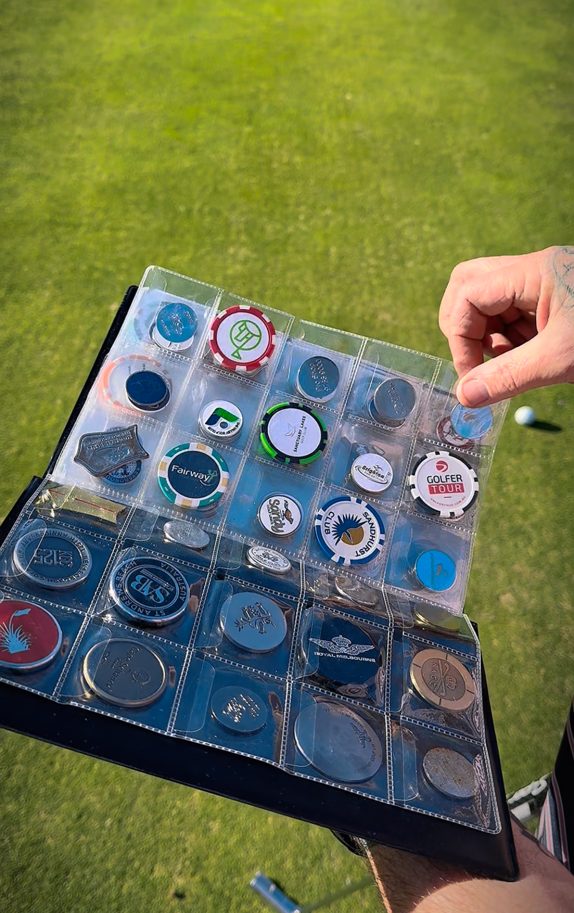A person's hand holding a collection of poker chips in a plastic display case at a golf course. The background is a well-maintained grass field.