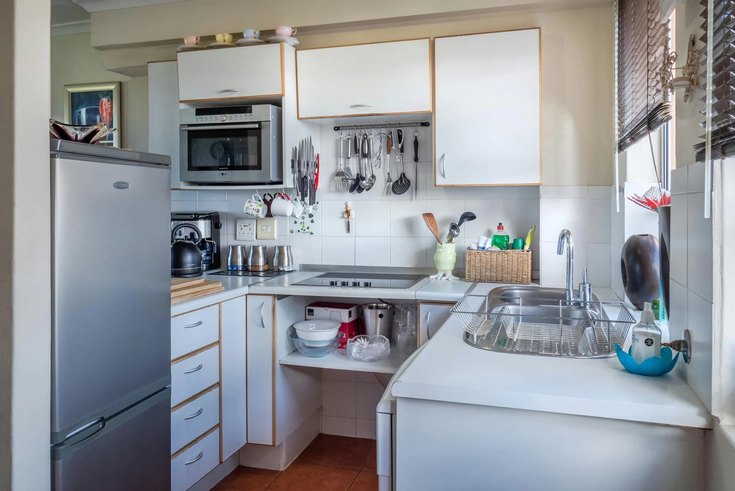 Compact kitchen with white cabinets, stainless steel refrigerator, microwave, and countertop appliances. Wall-mounted hooks hold knives and utensils, and there is a window with blinds over the sink.