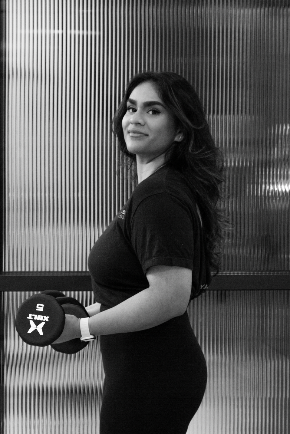 A woman with long, wavy hair holding a dumbbell and looking at the camera in a gym.