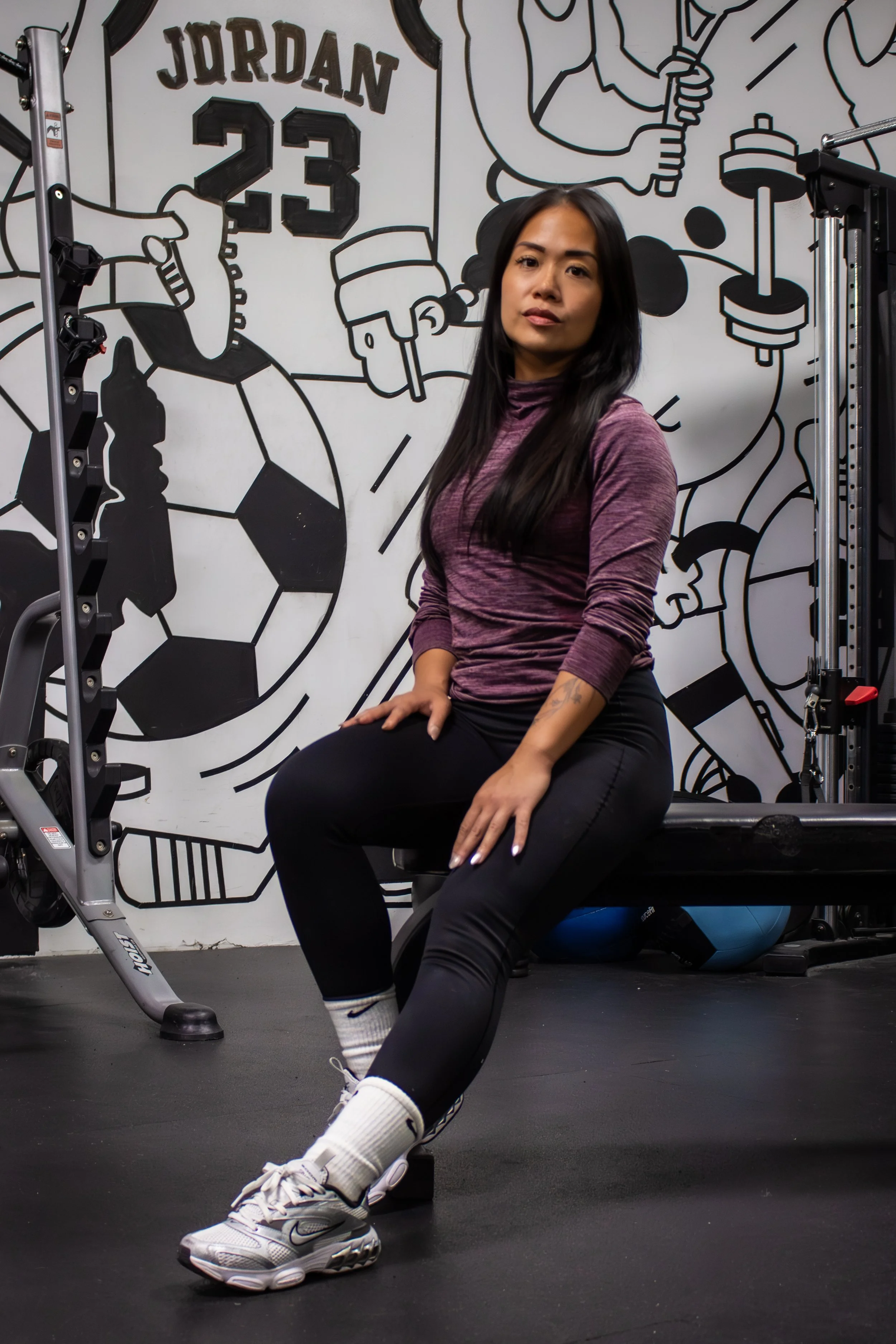 A woman in workout clothes sitting on a bench in a gym with a black and white mural of sports elements and the name 'Jordan' with the number 23 on the wall behind her.