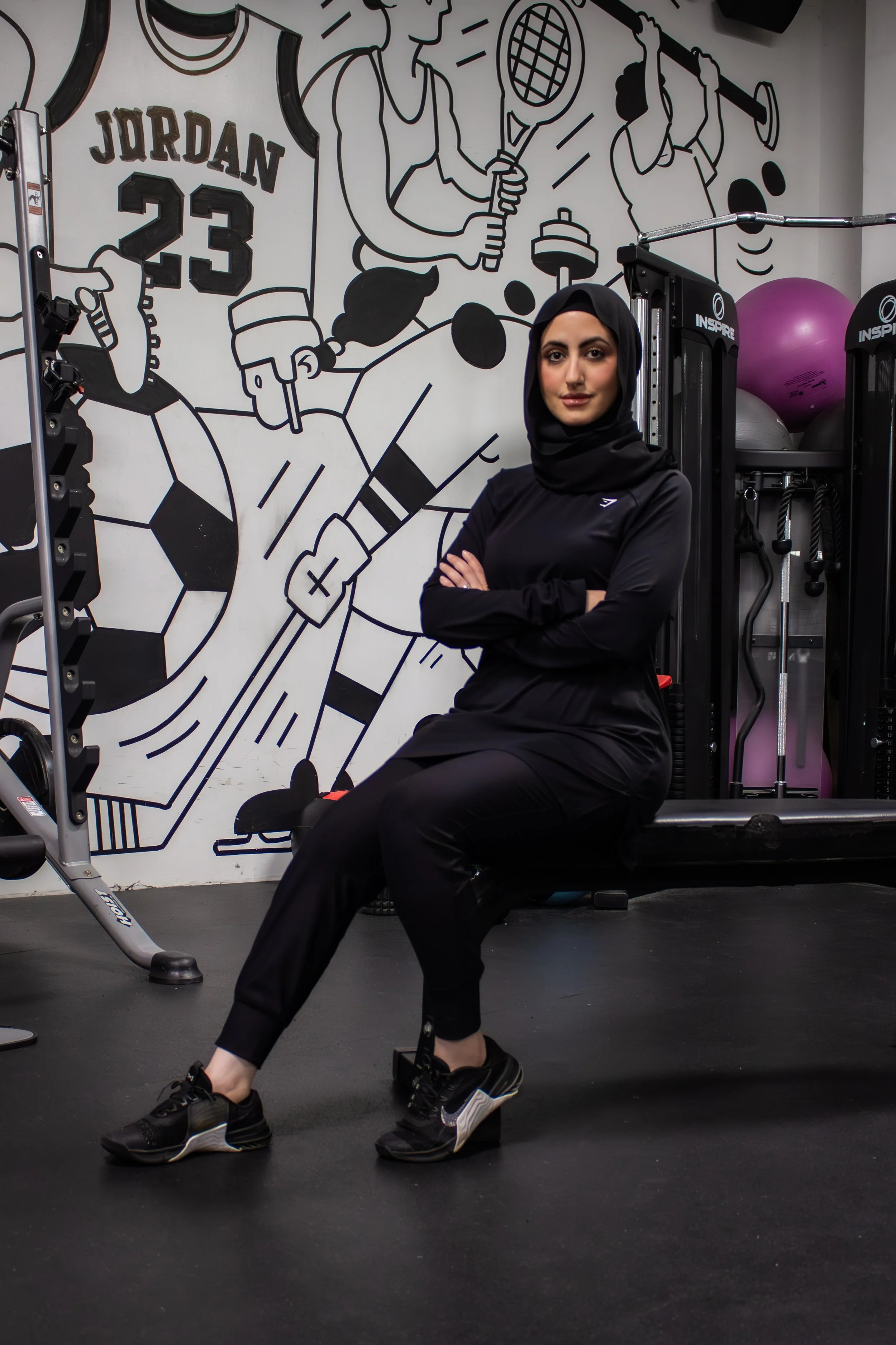 A woman wearing black sportswear and sneakers sitting on the floor of a gym, crossed arms, with a gym wall art showing athletes and sports equipment in the background.
