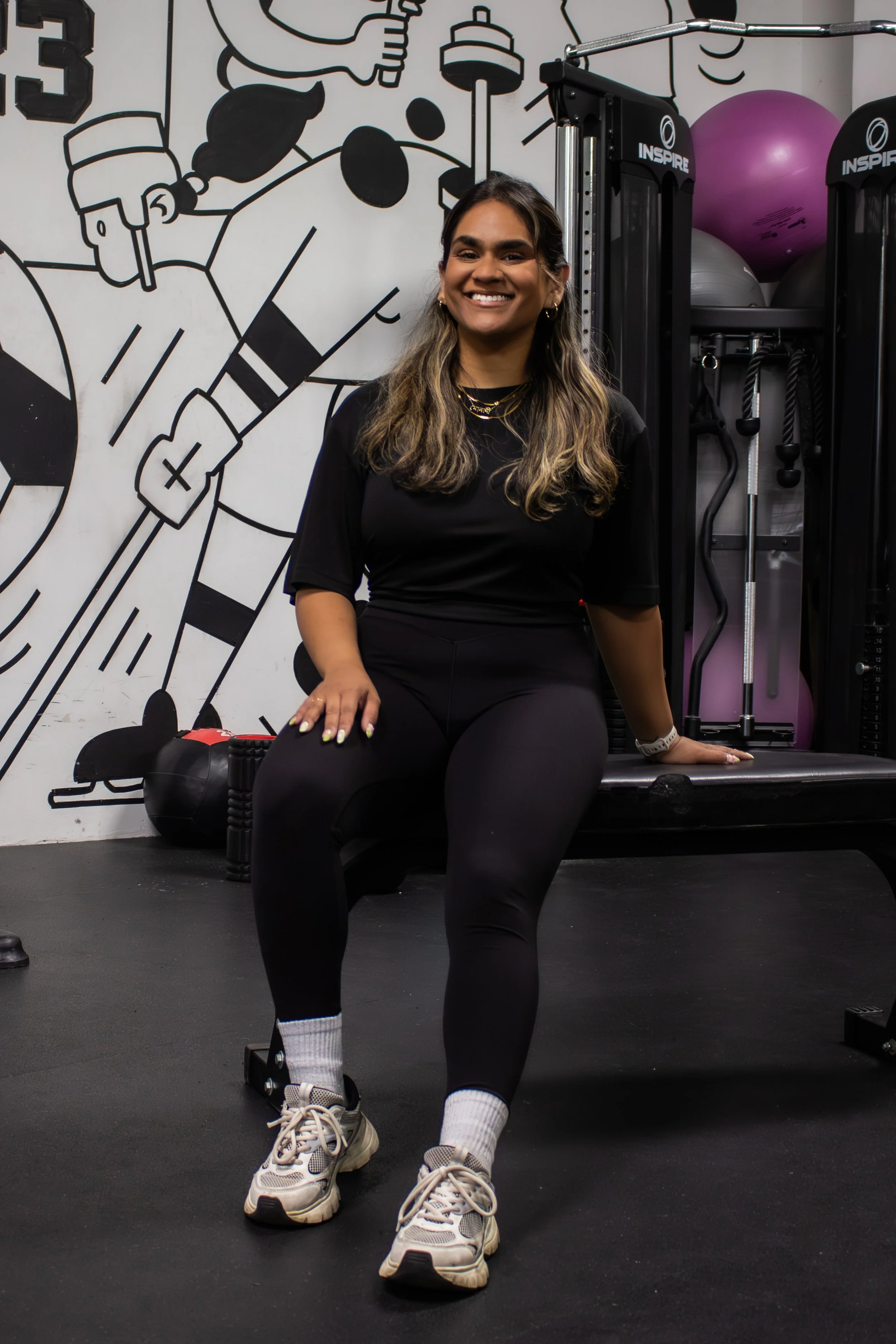 A woman sitting on a workout step in a gym, smiling at the camera. She is wearing black workout clothes, white sneakers, and white socks. Behind her is a wall with a black and white cartoon-style mural and exercise equipment, including purple and gray stability balls.