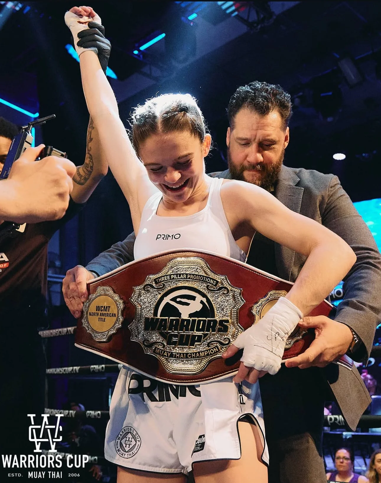 A female Muay Thai fighter is celebrating her victory holding a championship belt that reads 'Warriors Cup' and 'Muay Thai Champion'. She is smiling, and her right arm is raised while a man in a suit supports her from behind. She is wearing boxing gloves, a white tank top with the logo 'PRIMO', and Muay Thai shorts. The background shows a boxing ring with bright lights and spectators.